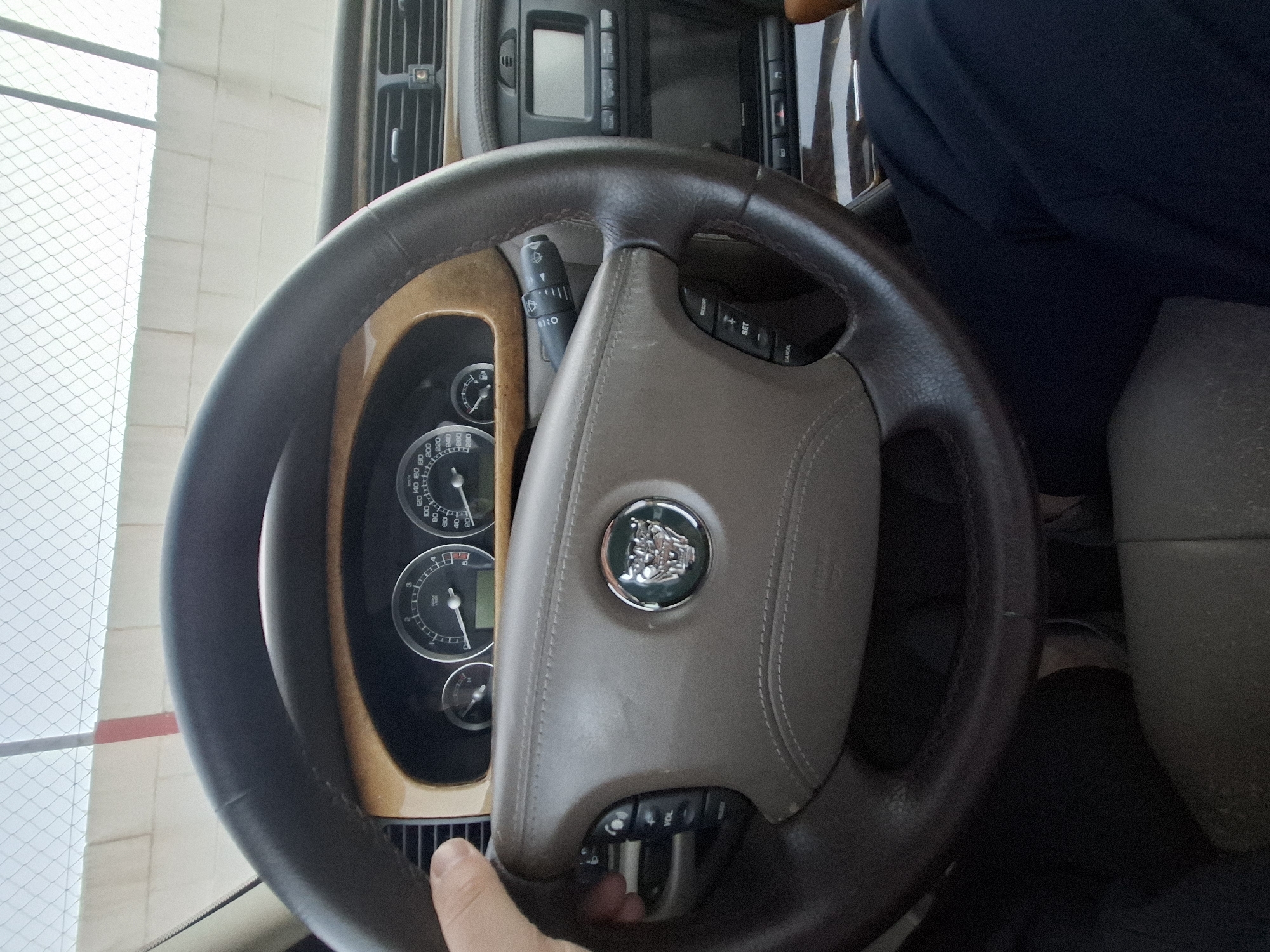 A close-up shot captures the steering wheel and dashboard of a Jaguar car. The steering wheel is dark brown leather, with a light brown leather center pad emblazoned with the Jaguar emblem. The dashboard features analog gauges displaying speed and other vehicle information, surrounded by wood trim. A control panel with various buttons is visible above the steering wheel. A person wearing a navy blue shirt is partially visible in the right side of the image. The environment appears to be inside a car, and the location is unknown.