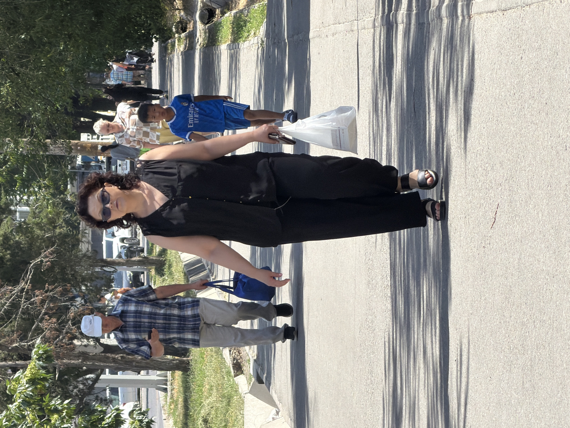 The image shows a street scene with several pedestrians walking on a sidewalk on a sunny day. In the foreground, a woman with dark, curly hair and sunglasses is walking towards the camera, carrying a white plastic bag in her right hand and a phone in her left hand. She is wearing a black sleeveless top and black pants. Behind her, a young boy in a blue t-shirt with "Emirates Fly Better" printed on it and blue shorts is walking away from the camera. Further back, a man in a plaid shirt and light-colored pants is walking away from the camera, holding a blue bag and looking at his phone. Other pedestrians and trees are visible in the background. The sidewalk is paved, and a street with cars is to the left. The environment suggests an urban setting.