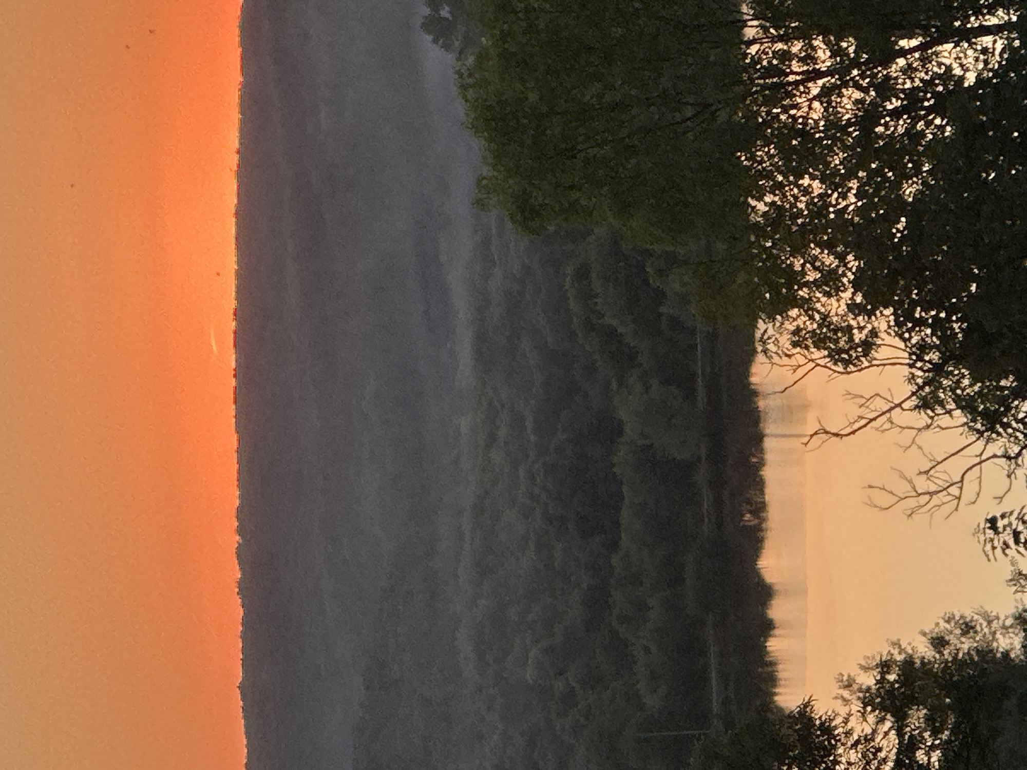 This is a photograph of a landscape at sunset, with the sky a vibrant orange hue. In the foreground, silhouetted trees with bare branches frame the scene, with denser foliage to the right. Below the horizon, a dark, undulating line of trees stretches across the middle ground, partially obscuring a body of water. The water reflects the warm colors of the sunset, creating a shimmering surface. The overall impression is one of a peaceful, natural setting, likely in a rural or undeveloped area. The location in the city of Makiv, Ukraine, suggests a possible setting within the Ukrainian countryside.