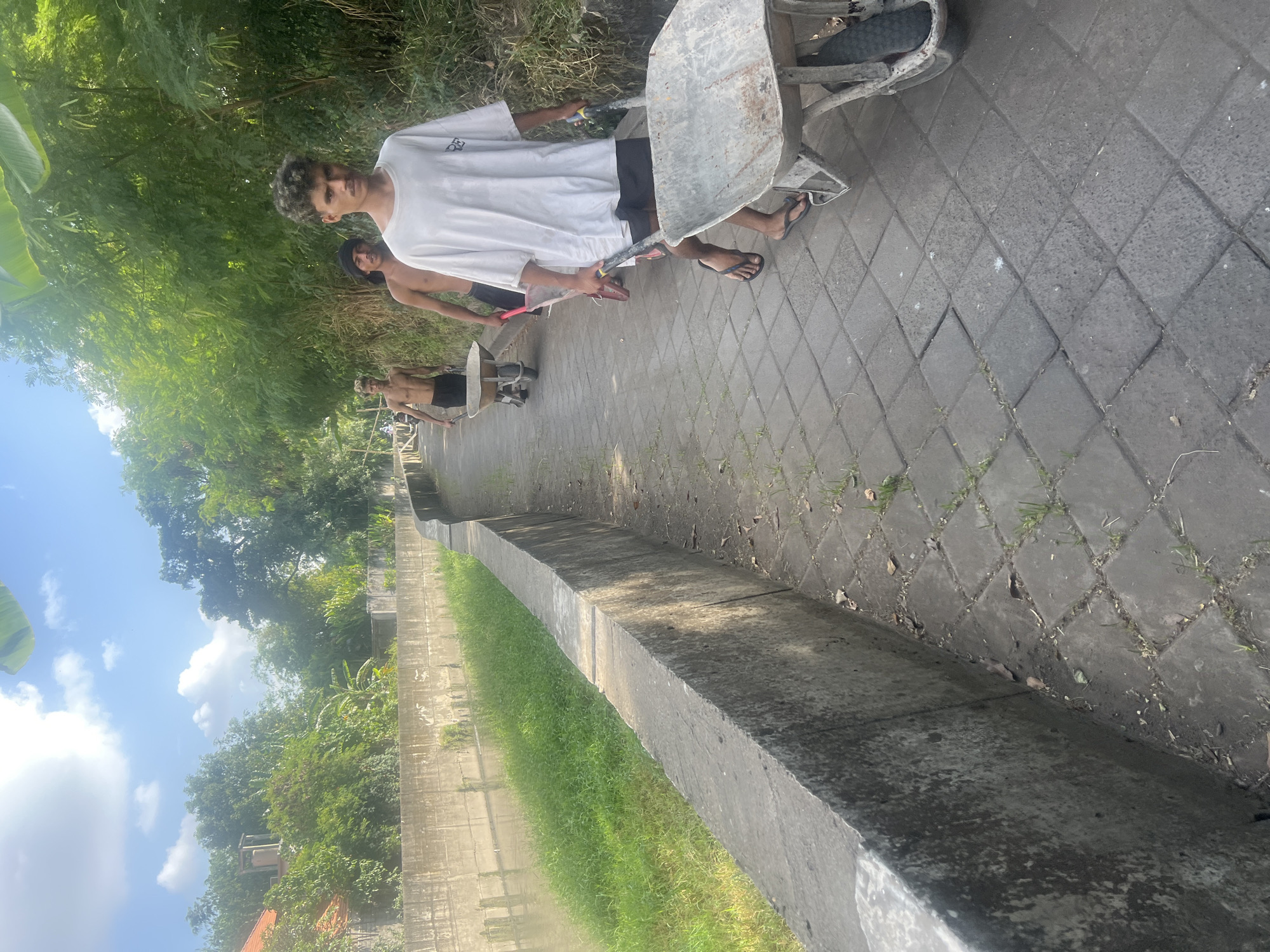 In the city of Denpasar, Indonesia, three young men are walking along a paved pathway next to a concrete drainage canal. The canal has a narrow strip of green grass growing along its edge. The pathway is bordered by lush vegetation, including trees and bamboo. The sky above is bright blue with scattered white clouds.
The man in the foreground, wearing a white t-shirt and dark shorts, is pushing a wheelbarrow filled with what appears to be dirt or debris. He is wearing flip-flops. Behind him, a second man, shirtless and wearing dark shorts, is also pushing a wheelbarrow. He has a black cloth wrapped around his head. Further back, a third young man, shirtless and wearing dark shorts, is walking with an empty wheelbarrow. All three men appear to be engaged in manual labor, likely cleaning or maintaining the canal and its surroundings. The overall scene suggests a warm, sunny day in a tropical environment.