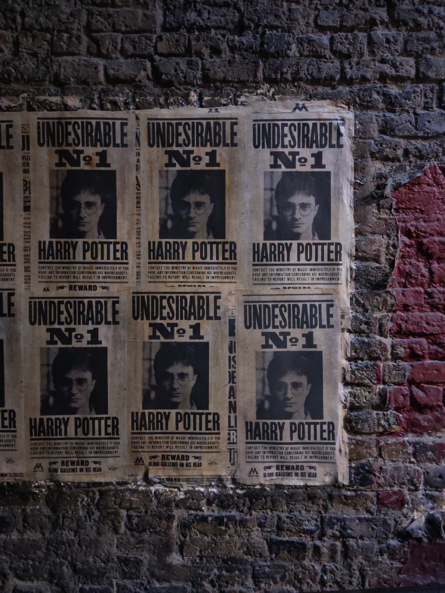 The image depicts a brick wall covered with "Undesirable No. 1" posters featuring Harry Potter. The posters are aged and slightly torn. The brick wall appears to be part of a building with some red paint visible on the bricks. The posters offer a reward of 10,000 Galleons for information leading to Harry Potter's whereabouts. The location is in Watford, United Kingdom.