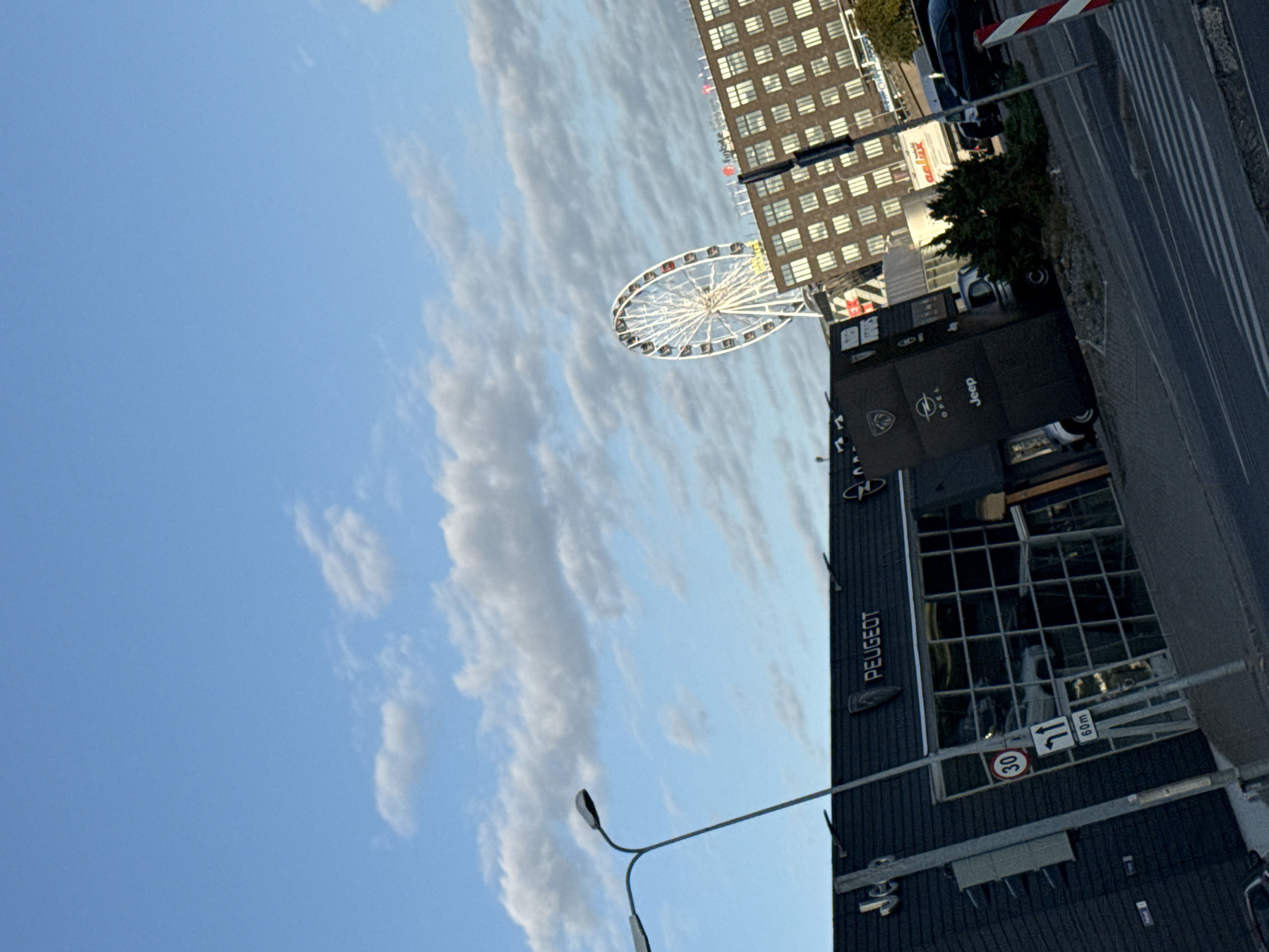 The image captures a street view in Tallinn, Estonia, with a slight tilt to the right. In the foreground, a street lamp is visible, casting a long shadow. To the left, a dark building with "PEUGEOT" and "OPEL" signage is apparent, featuring large glass windows. A traffic sign indicating a 30 km/h speed limit and an arrow pointing straight ahead with "60m" text is also present. Further in the background, a Ferris wheel stands tall against a partly cloudy blue sky. A multi-story building with numerous windows dominates the right side of the frame, with a sign that appears to read "finbite". The street in front of the buildings is paved, with a pedestrian crossing marked by white stripes. A few cars are parked or driving on the street. The overall scene suggests an urban environment with commercial establishments and recreational attractions.