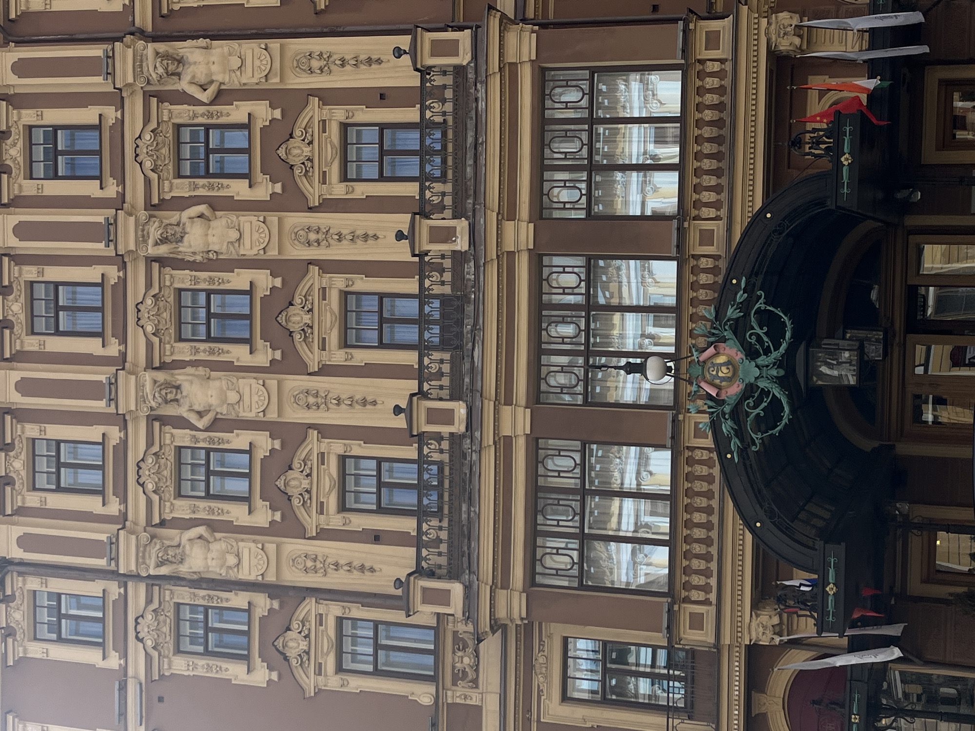 The image shows a section of a building facade in Saint Petersburg, Russia. The building is multi-storied with a facade of brown and beige. The facade features a pattern of windows, with decorative window surrounds, including figures of reclining men. Between the window sections are decorative moldings. To the right of the building is an archway with decorative foliage on the sides. The photo is taken from below, pointing up.