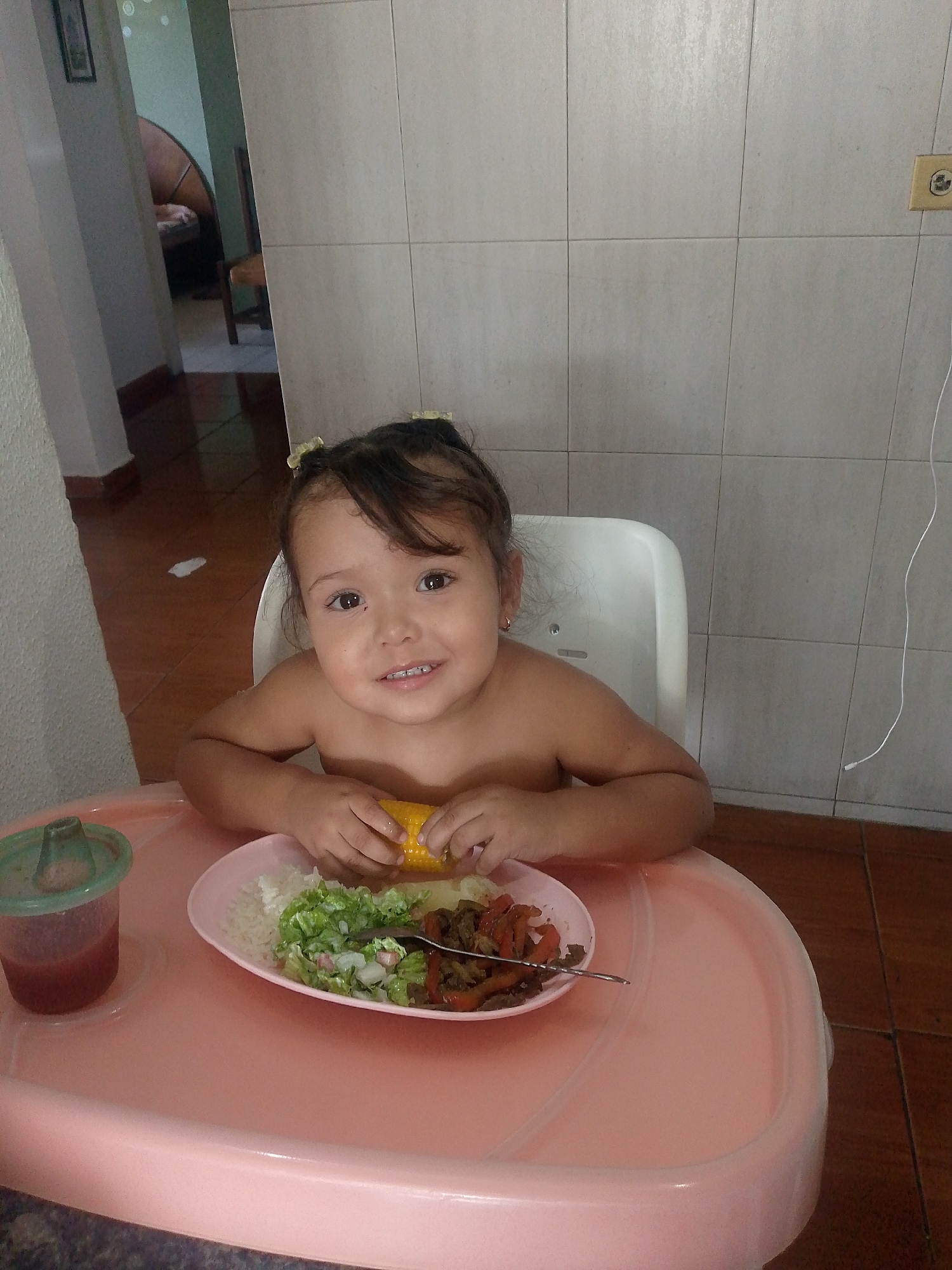 A young child sits at a pink highchair, positioned in a room with tile walls. The child, with dark hair and brown eyes, is holding a cob of corn. A plate on the highchair contains rice, salad, and some cooked vegetables and meat. A covered cup with a red liquid is also on the tray. An electrical outlet is visible on the tiled wall behind the child. A doorway is seen in the background, showing a glimpse into another room with furniture. The location is Caracas, Venezuela.
