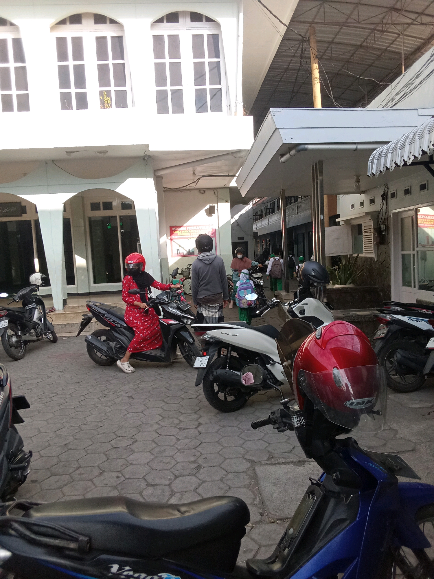 In Jember Kidul, Indonesia, a street scene unfolds with multiple motorcycles parked on a cobblestone surface in front of a white building with arched windows. A woman in a red floral dress and a red helmet is seated on a black motorcycle, while a person in a grey hooded shirt and a patterned sarong stands nearby. Other motorcycles are visible, some with riders and some parked. The environment suggests an outdoor setting, possibly near a school or community center, with a covered walkway and visible utility lines overhead.