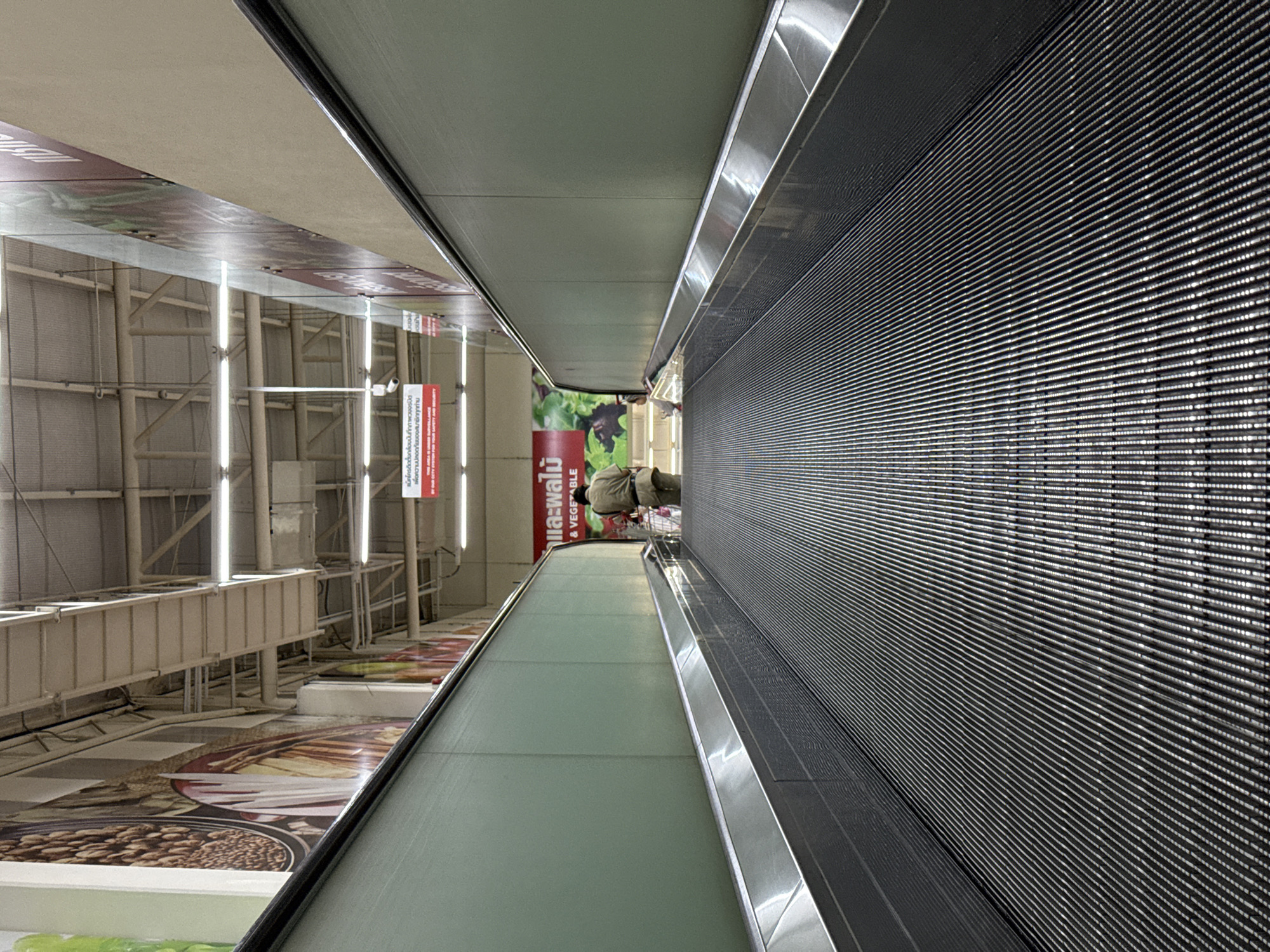 This image depicts a moving walkway in a shopping mall or supermarket in Bangkok, Thailand. A person is visible on the walkway, pushing a shopping cart. Adjacent to the walkway is an advertisement featuring food and produce. The architectural structure of the building is visible, including the ceiling and overhead lighting.