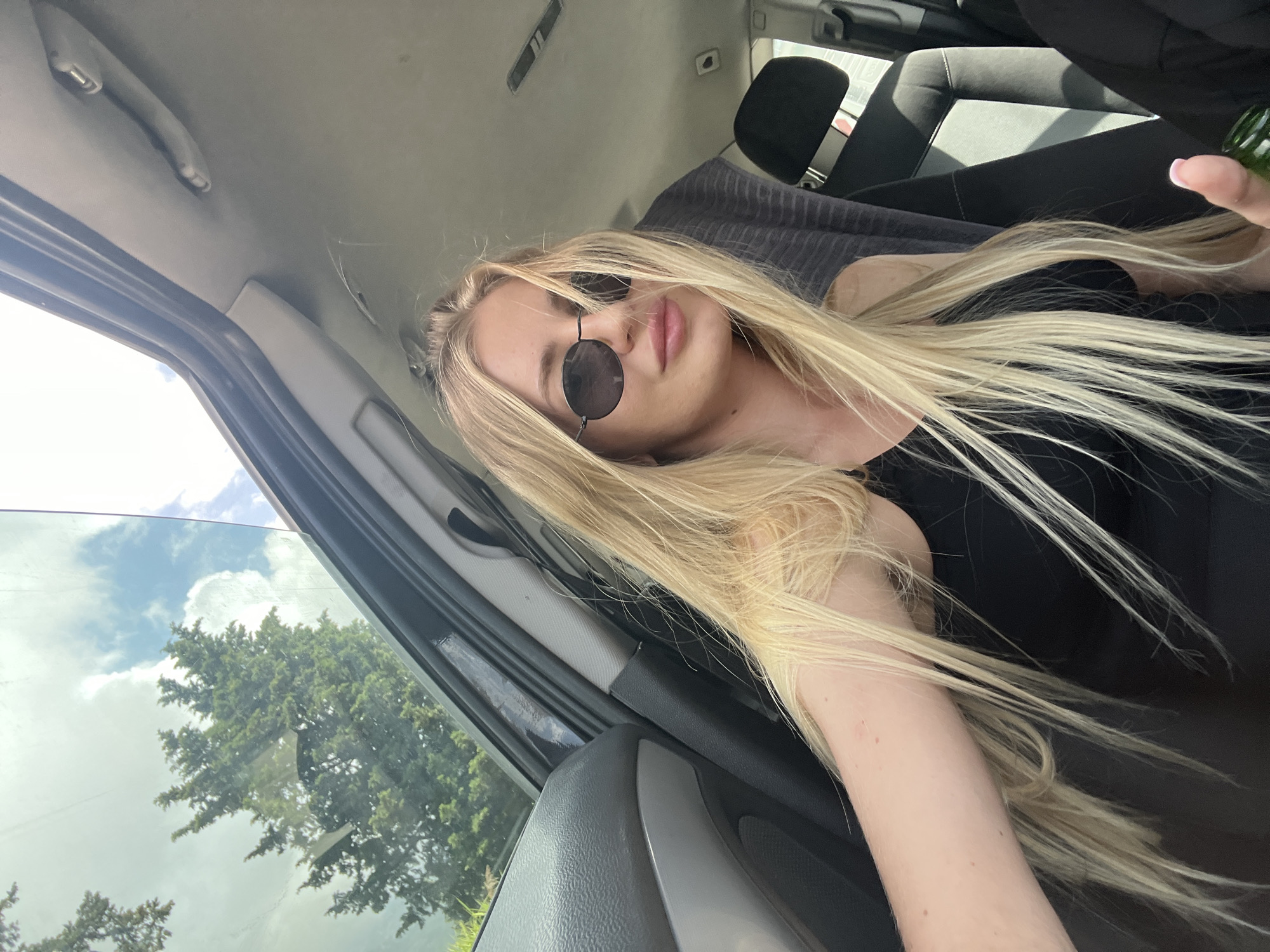 A selfie-style photograph, taken from within a car, shows a blonde woman in the front passenger seat. She is wearing round sunglasses and a black top. Her blonde hair is long and straight. The car's interior is visible, with the dashboard and passenger seat in view. Through the open car window, green trees and a cloudy sky are visible. The location is consistent with an area around Budva, Montenegro, considering the observed Mediterranean vegetation.