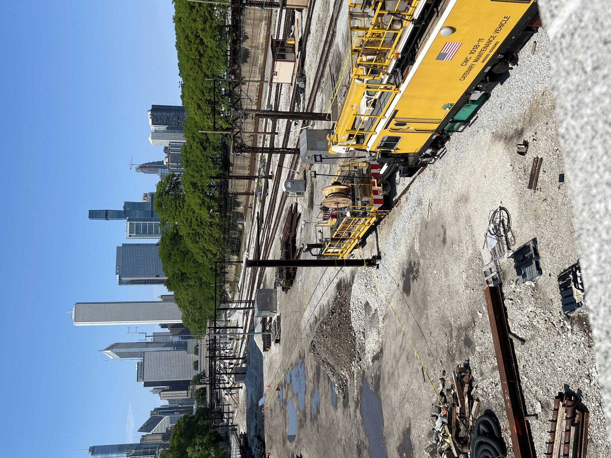 A high-angle, daytime shot shows a construction and maintenance area alongside multiple train tracks, with the cityscape of Chicago, Illinois in the background. A yellow Catenary Maintenance Vehicle with the American flag and the text "CWC 1018-11 CATENARY MAINTENANCE VEHICLE" is visible on the right side of the frame, partially on a gravel surface. Multiple train tracks extend toward the city skyline, parallel to a line of green trees. The maintenance area has puddles, construction materials, and debris scattered around. Buildings of various heights are visible in the cityscape, including the John Hancock Building, one of the city’s famous skyscrapers. Overhead power lines and train signal equipment run alongside the tracks. The sky is clear and blue.