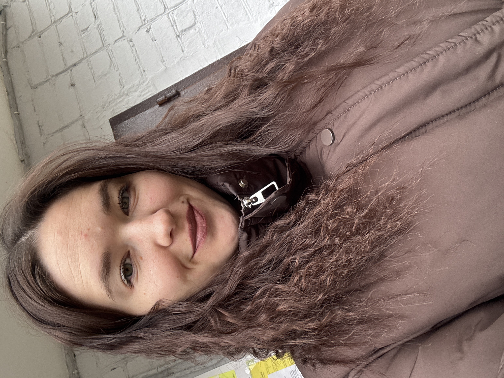 A woman with brown curly hair, wearing a brown jacket, is facing the camera. She is smiling slightly. Behind her is a white brick wall. The location is Chaikovsky, Russia.
