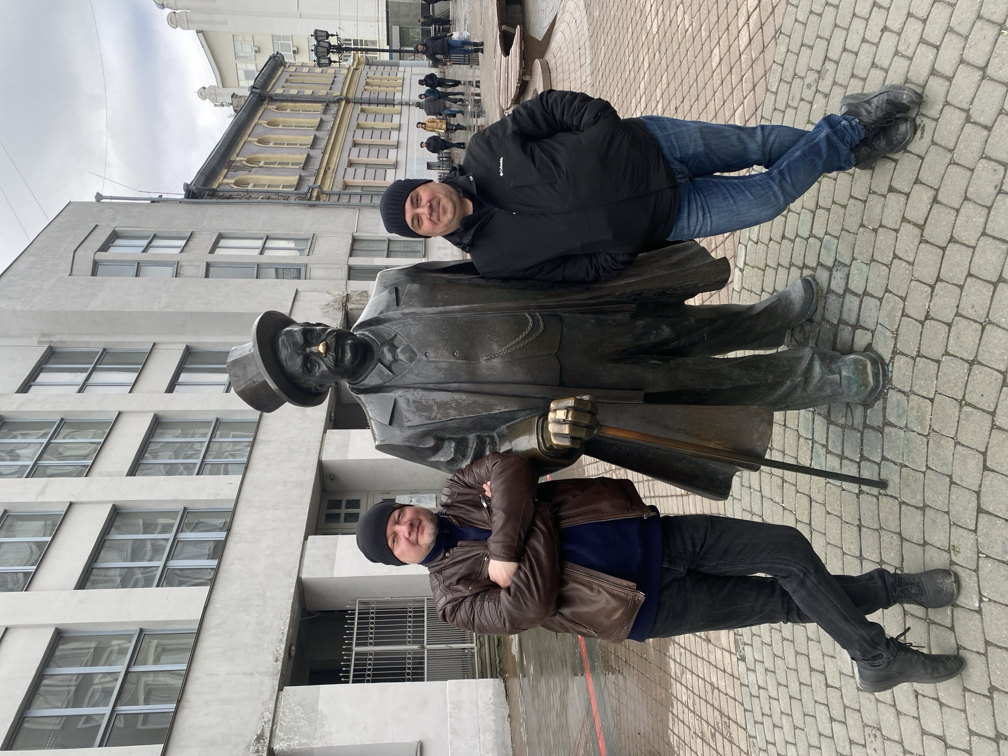 Two men pose with a bronze statue of a man in a suit and hat, likely a public art installation. The statue has a gold tooth. One man wears a brown leather jacket and a black beanie, and he stands with his arms crossed next to the statue. The other man wears a black winter coat and beanie, and leans against the statue. The location appears to be Yekaterinburg, Russia, judging by the architecture and brick paving. In the background, other pedestrians walk down the street, and buildings with windows are visible. The sky appears overcast.
