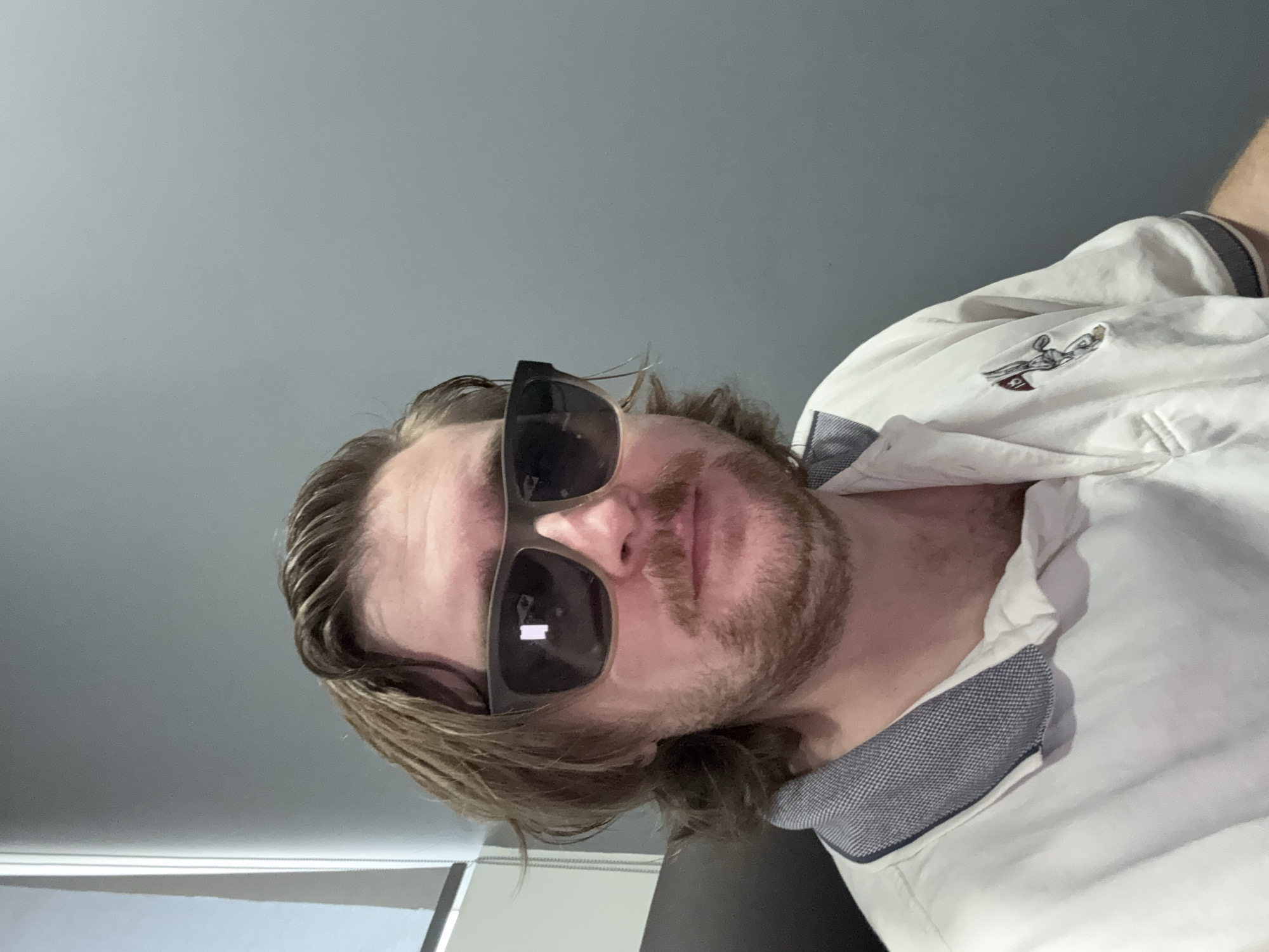 A man with long blonde hair and a reddish beard wears sunglasses and a white polo shirt with a gray and blue patterned collar. He is positioned indoors against a gray background. The polo shirt has a small logo of a figure holding a golf club on the upper left side. No location information about Cuautitlán Izcalli, Mexico, is discernible from the image.