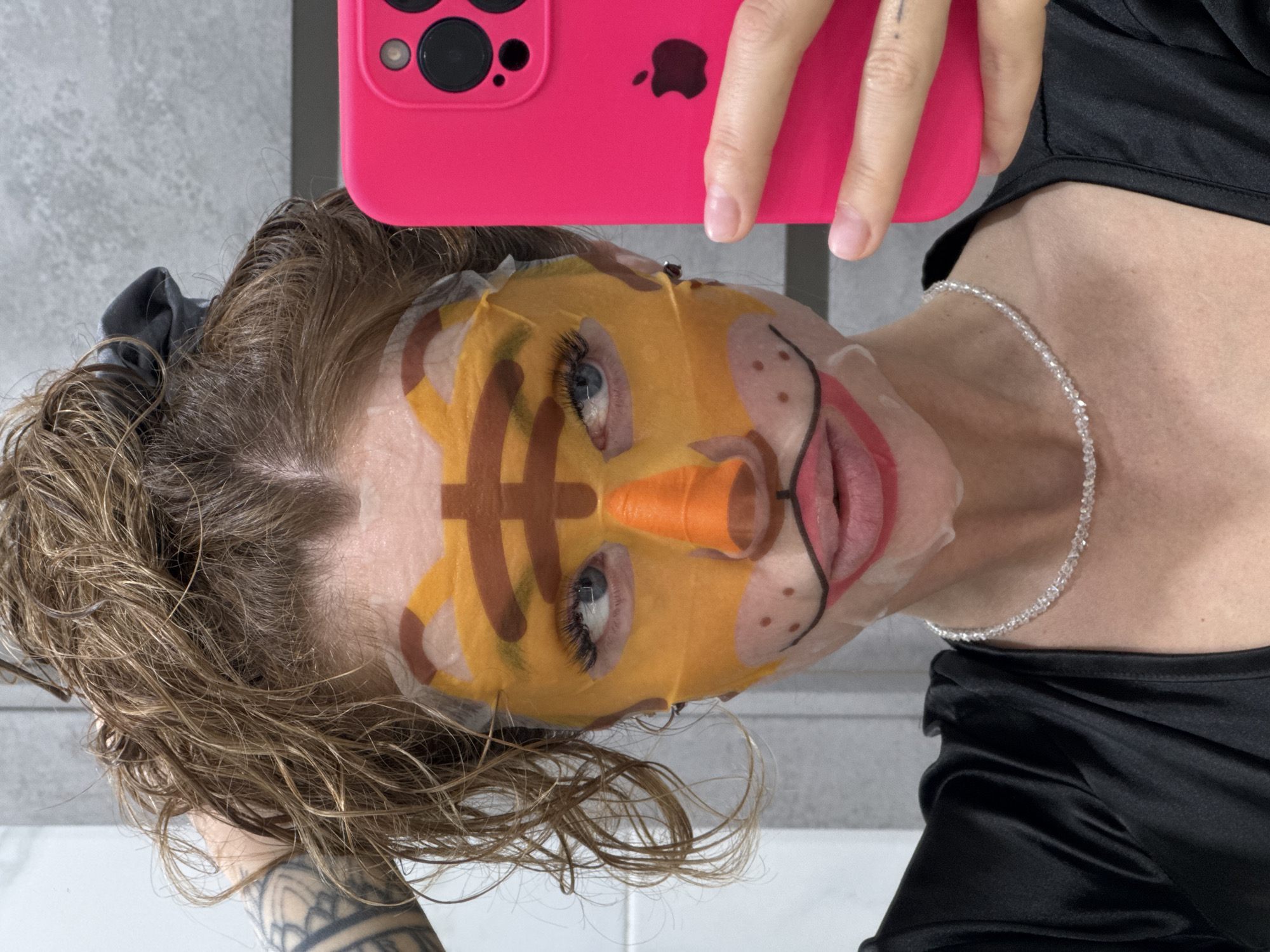 A person is taking a selfie with a pink iPhone while wearing a facial mask designed to resemble a cartoon character, likely indoors. The person has blue eyes, light brown hair, and is wearing a necklace. Their arm and a tattoo are visible in the lower left corner. The person is wearing a black top. The background is a tiled wall, and a sink surface is visible in the bottom left corner. No evidence suggests this image was taken in the city of Chaikovsky, Russia.