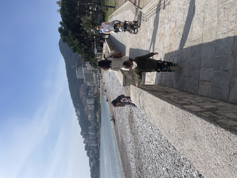 The image displays a sunlit coastal scene in Budva, Montenegro, specifically along a promenade adjacent to a pebble beach. In the immediate foreground, a light-colored flagstone promenade runs parallel to a low, rough-hewn stone wall separating it from the beach.
A young boy, approximately 4-6 years old, in a dark green hooded top and multi-colored patterned trousers, walks away from the camera on the promenade, extending his right hand to touch the stone wall. Directly in front of him, a woman, seen from behind, walks along the promenade, wearing a light-colored long coat or cardigan and carrying a brown shoulder bag. To the left, partially obscured by the stone wall, a young girl in a colorful patterned jacket stands on the beach side of the wall, facing towards the water. Further along the promenade, a woman in a white t-shirt pushes a three-wheeled stroller containing an infant or toddler.
The wide pebble beach, consisting of light grey and white stones, extends into the calm, clear, light blue-green waters of the Adriatic Sea. Several individuals are visible scattered across the beach in the midground, some seated directly on the pebbles, others walking.
The background features the city of Budva, characterized by numerous modern, multi-story buildings and high-rises lining the coastline and ascending the lower slopes of steep, tree-covered hills. One prominent hill, topped with a communication tower, is visible on the right. The sky is clear and light blue, with minimal atmospheric haze. Long shadows are cast across the promenade from objects and individuals, indicating direct sunlight from a high angle.