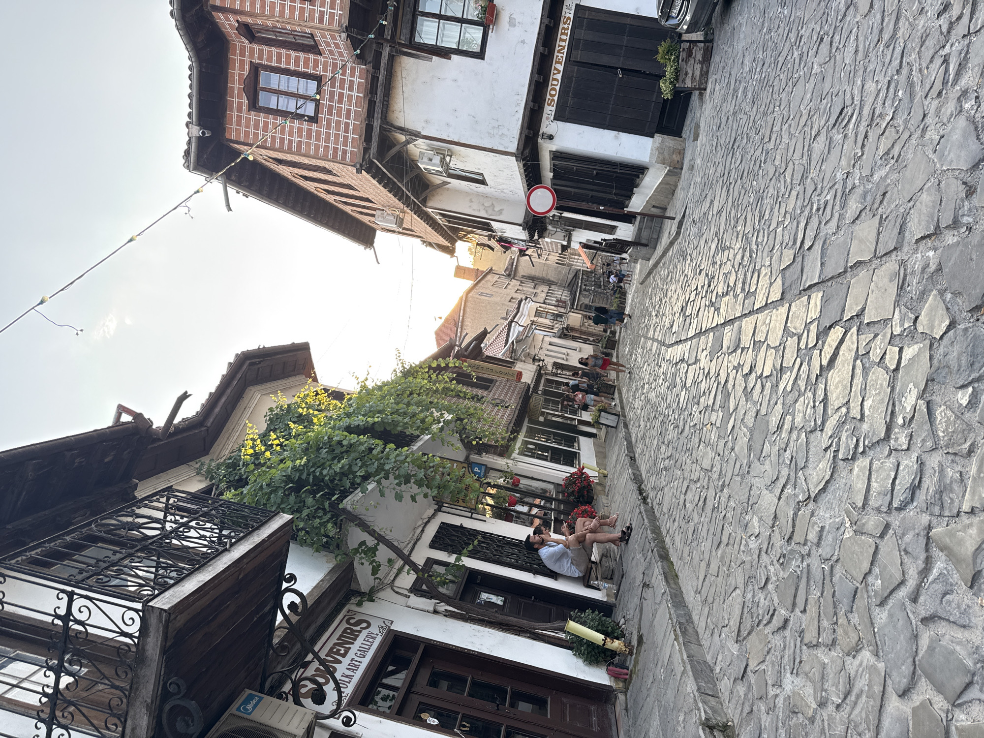 The image depicts a cobblestone street in Veliko Tarnovo, Bulgaria, likely in the historic Old Town area, characterized by traditional Revival-era architecture. The street is paved with irregular, flat stones, worn smooth by time. Buildings lining the street exhibit white plaster walls with dark wooden window frames and shutters, and some have ornate wrought-iron balconies. One building on the left prominently displays a sign reading "SOUVENIRS FOLK ART GALLERY." Green ivy cascades down the facade of this building, adding a touch of nature.
Several people are present. On the left side of the street, a couple is seated on a wooden bench, appearing to converse. Further down the street, a small group of people are walking, their figures silhouetted against the bright sky. Another individual is walking away from the camera on the right side. The sky is bright, suggesting it is either morning or late afternoon, with a warm, natural light illuminating the scene. String lights are visible stretched across the street, indicating a festive or evening ambiance. The overall impression is one of a charming, historic European street scene.
