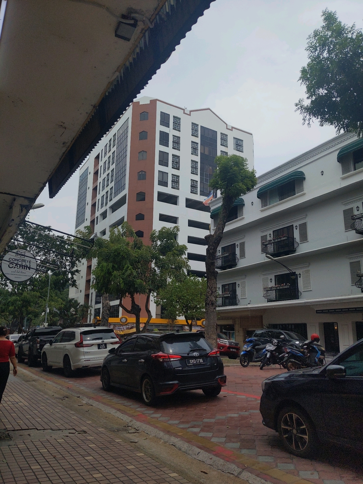 The image shows a street scene in Kota Kinabalu, Malaysia. In the foreground, a sidewalk with patterned paving stones is visible, with several cars parked along the street. A woman in a red shirt and black pants is walking on the sidewalk. The parked cars include a black Toyota sedan, a white SUV, and other vehicles further down the street. A group of motorcycles is also parked on the street.
Behind the cars, two buildings are prominent. The building on the left is a tall, multi-story structure with a white facade and brown accents. It has numerous windows, some of which are dark. A circular sign with the text "CONCRETE JUNGLE CABIN" is visible hanging from a pole on the left side of the image. The building on the right is a lower, white structure with green awnings over the windows. Balconies with decorative railings are visible on this building.
Trees line the street and are interspersed between the buildings, suggesting an urban environment with green spaces. The sky is overcast. The overall impression is of a busy street in a city, likely during daylight hours, given the ambient light.