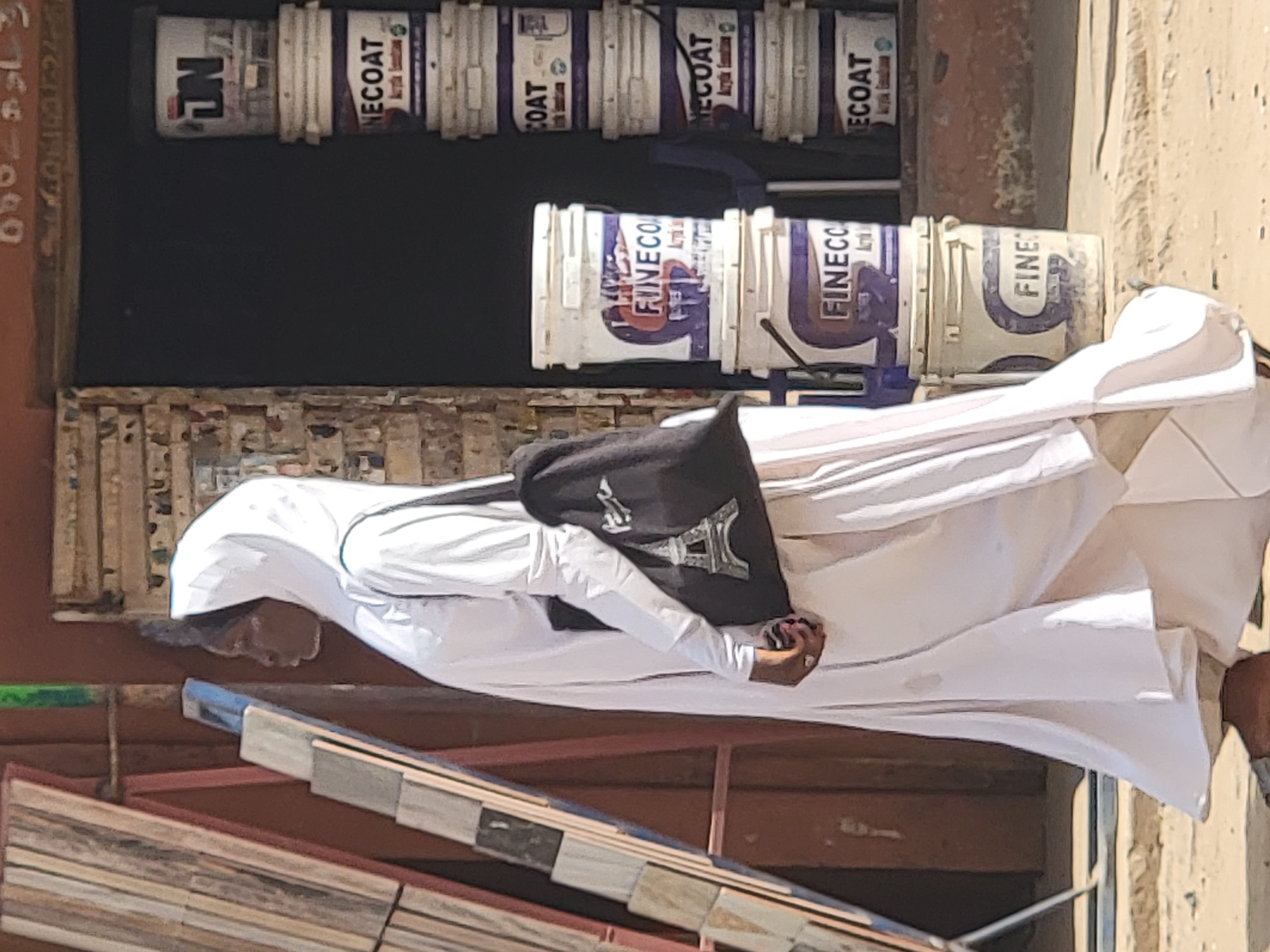 A person wearing a long white robe with a black bag is walking past a store in Maiburiji, Nigeria. The store sells paint, including "Fine Coat" and "FinecCo" brands. The person's head is covered and they are carrying a black bag with the Eiffel Tower on it.