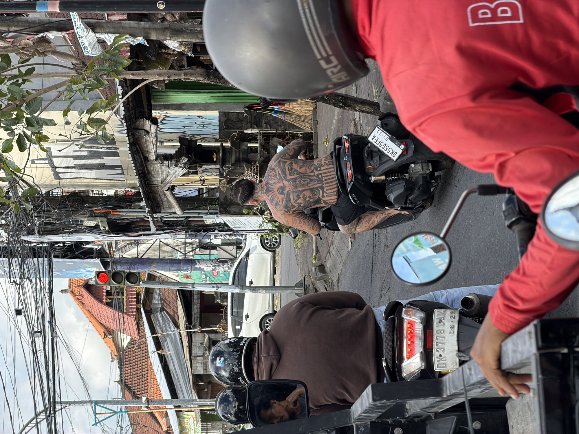 A bustling street scene in North Kuta, Indonesia, captures several people on scooters stopped at a red traffic light during daylight hours. The image is taken from the perspective of someone riding a scooter, with their grey helmet (bearing "ARC" text) and red long-sleeved shirt (with a white "B" logo) partially visible in the foreground.
Immediately in front, a person wearing a brown hooded sweatshirt and a black helmet is also stopped on a scooter. Their reflection, showing a man's face, is visible in the right-side mirror of the foreground scooter. The license plate of their scooter, "DK-3778 UAK" with "08-28" below it, is clearly visible.
Further ahead, a man with extensive, intricate black tattoos covering his entire back and arms rides a black scooter, appearing shirtless and wearing black shorts. His hair is tied back. His scooter's license plate reads "DK5505FEA" with "01-31" underneath it, and "NAMX" is visible on the vehicle.
The background reveals a typical Indonesian streetscape with dense overhead utility wires crisscrossing the sky. Various buildings line the road, some with traditional tiled roofs and others with corrugated metal, appearing somewhat weathered. A bright red traffic light is visible among the wires, confirming the stop. A white car is parked further down the street, and a tree branch with green leaves extends into the frame from the left. The asphalt road surface indicates an urban or semi-urban setting, consistent with the specified location of North Kuta, Indonesia.
