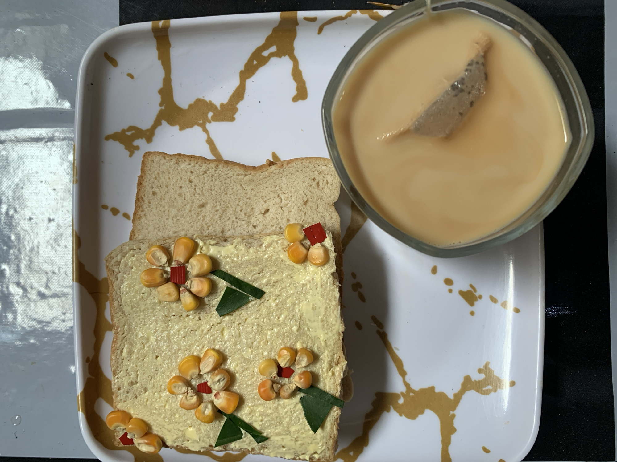The image shows a breakfast scene on a white plate with gold-colored splashes. On the left are two slices of bread, one plain and one topped with a yellow spread. The spread is decorated with what appear to be corn kernels and small red pieces, arranged in flower shapes, with green leaves as stems. To the right is a clear glass bowl filled with a light brown liquid, possibly tea or coffee, with a teabag visible inside. The plate is placed on a surface that is partly white and partly black. There is no evidence to indicate this image was taken in Rafin Par, Nigeria.