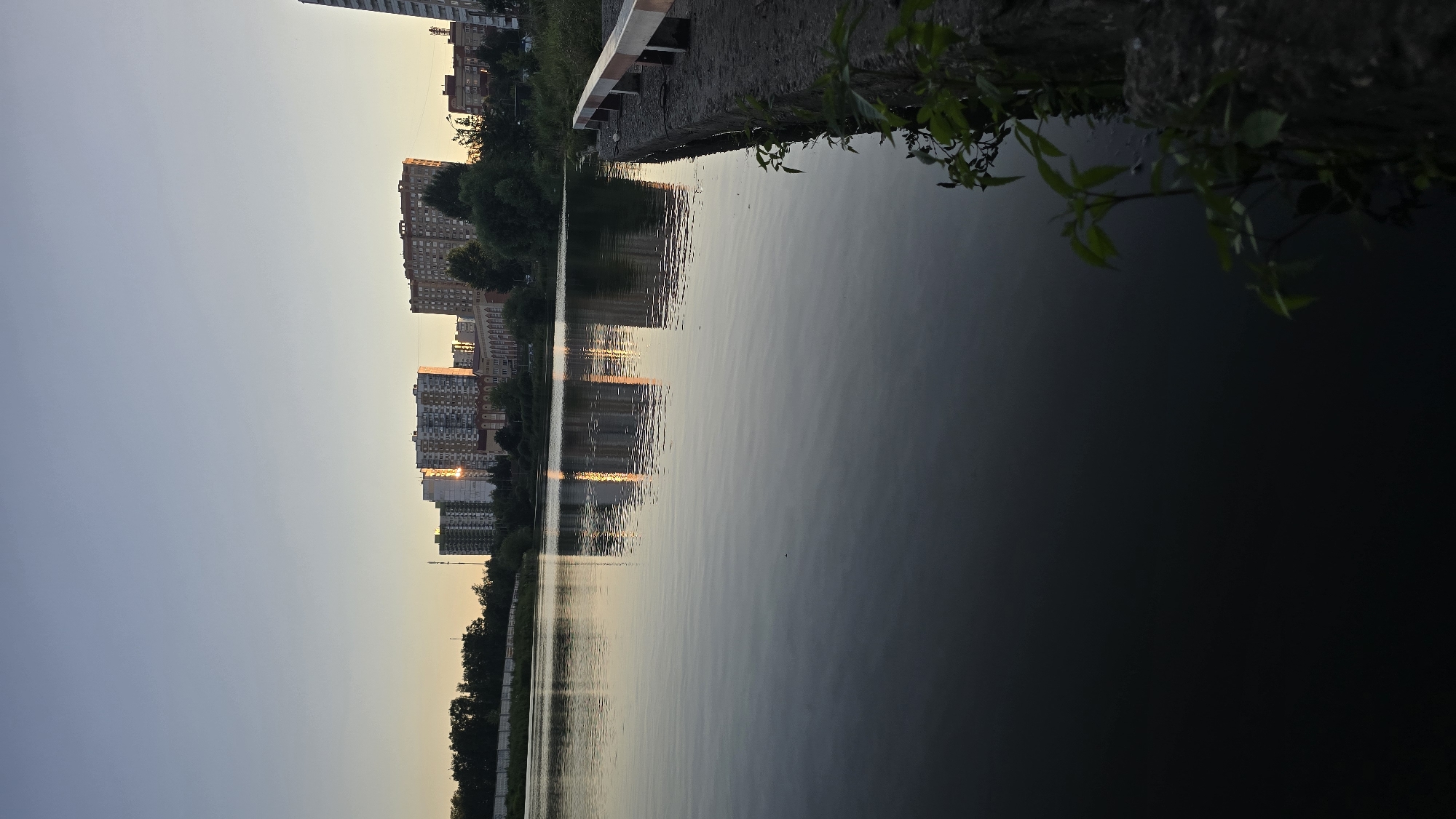The image shows a serene, waterfront scene at dusk in Ashcherino, Russia. The main subjects are a row of tall, residential apartment buildings lining the opposite bank of a calm body of water. The buildings are of varying heights and architectural styles, with many windows visible. Their reflections are clearly mirrored in the still water, creating a symmetrical and tranquil effect.
The activity taking place is a peaceful observation of the sunset. The time of day is evident from the soft, warm glow of the setting sun reflecting off the buildings and water, casting long shadows and illuminating the sky with hues of orange and yellow against a fading blue. The weather appears clear and calm, as indicated by the undisturbed surface of the water.
Notable details include the presence of trees and foliage along the embankment in the foreground, with some branches and leaves hanging over the water. The embankment itself is composed of a gravelly path and a white-and-red striped barrier. The overall mood conveyed is one of quietude and natural beauty, with the urban landscape softened by the twilight and the reflective qualities of the water. There is no visible text in the image.