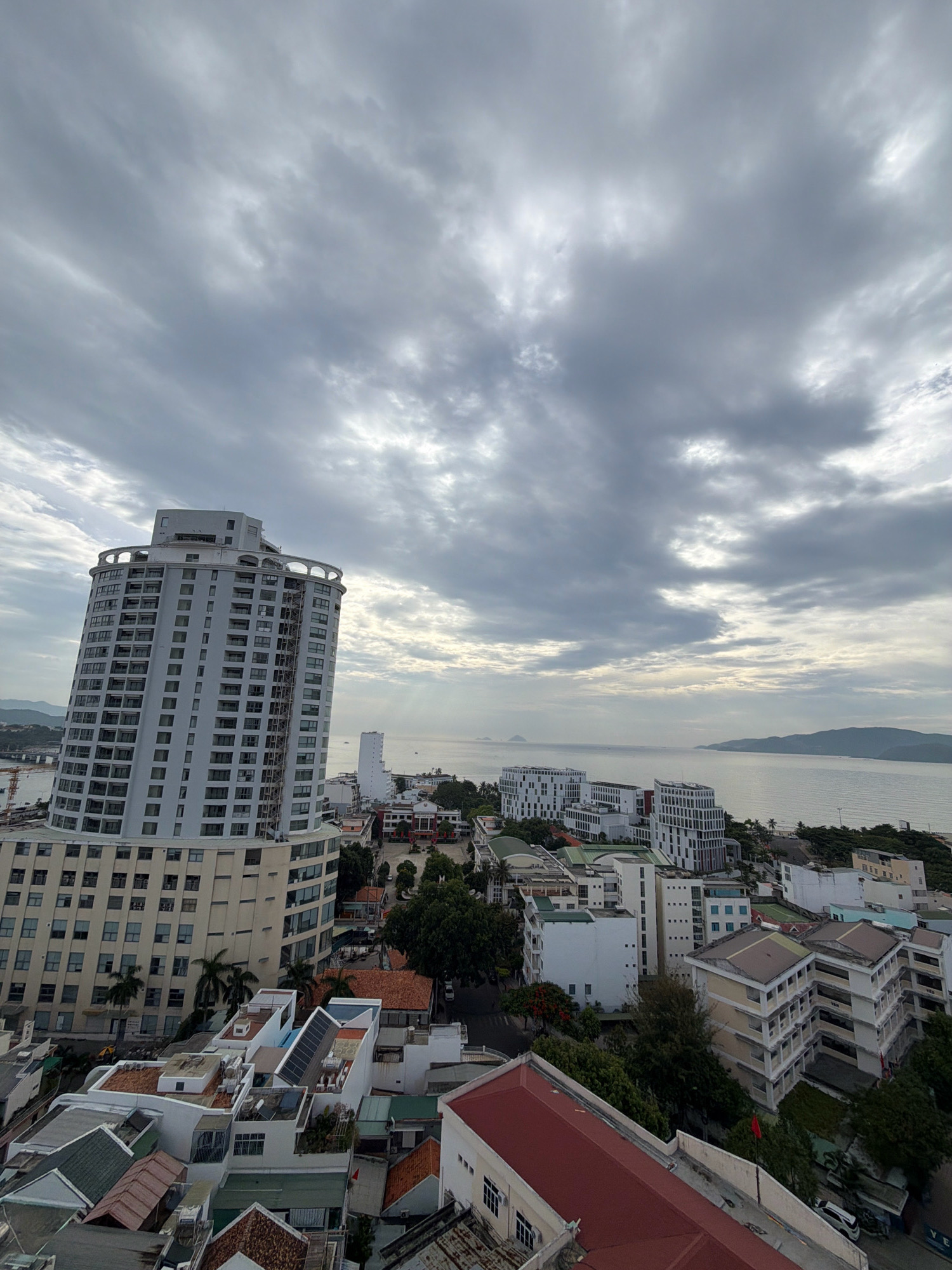The image provides an elevated, wide-angle view of a coastal city, likely Nha Trang, Vietnam, under a cloudy, overcast sky. In the foreground and midground, there are numerous buildings of varying sizes and architectural styles. A prominent, curved, multi-story building with a light-colored facade and numerous windows dominates the left side of the frame. Adjacent to it are smaller residential or commercial buildings with flat roofs, some featuring solar panels. Further into the city, a cluster of modern, white, multi-story buildings with repetitive window patterns are visible. Beyond the city buildings, a body of water, presumably the sea, stretches to the horizon. Hills or mountains form the backdrop on either side of the water. The sky is filled with dense, grey clouds, with some patches of brighter light suggesting the sun is diffused or attempting to break through. There are no visible people or vehicles in the immediate foreground or midground. The overall impression is a densely populated urban landscape meeting a coastline under a somber sky.