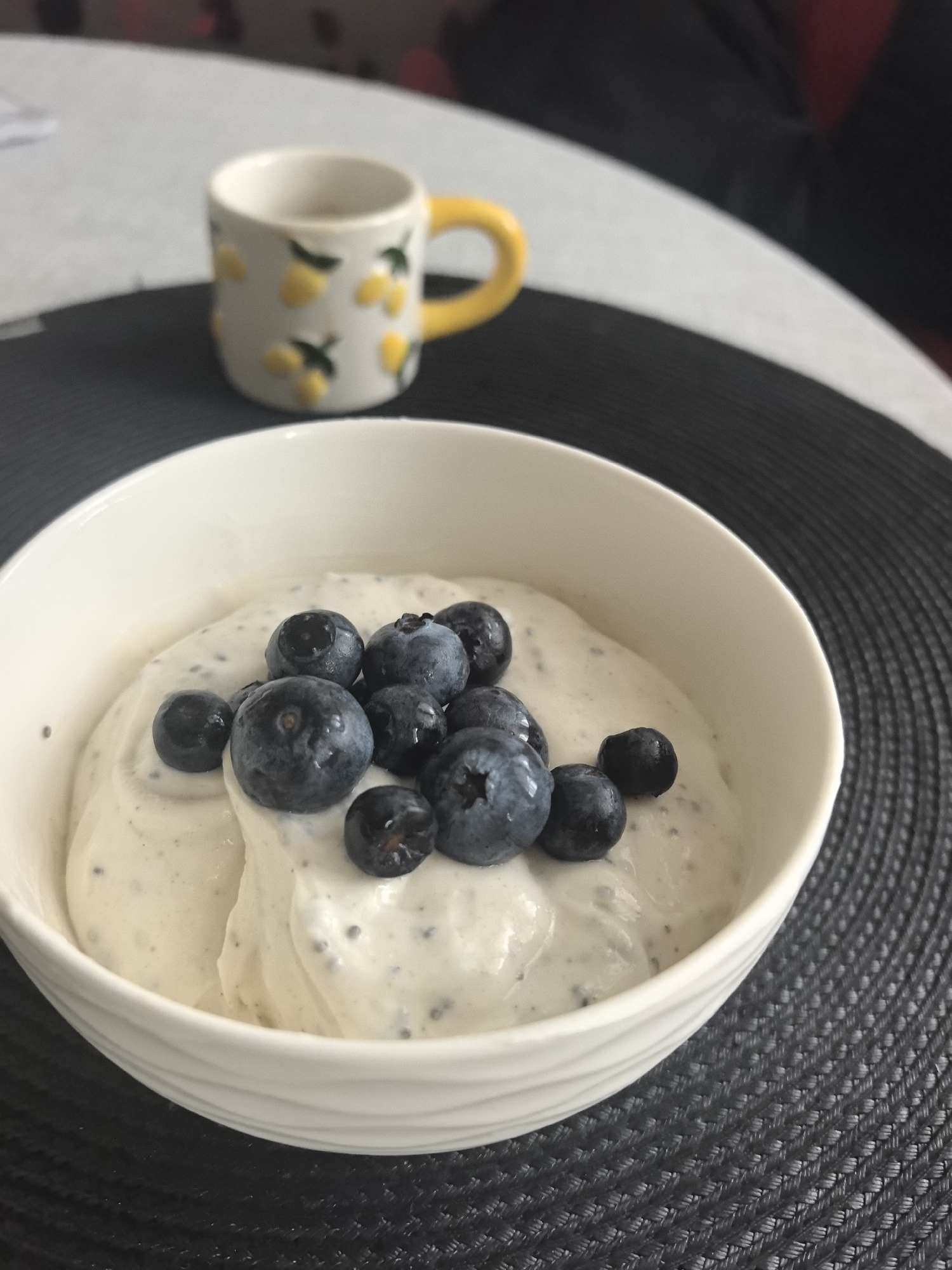 A close-up shot depicts a white bowl filled with a creamy, white substance speckled with small dark dots, likely chia seeds. The substance is topped with approximately ten fresh blueberries of varying sizes. Behind the bowl, a small ceramic cup with a yellow handle and a pattern of lemons is visible. The bowl and cup sit on a dark, woven circular placemat. The background is blurred, but a glimpse of a white surface and a red object suggest a table setting. The image does not contain any information to indicate it was taken in Stara Zagora, Bulgaria.