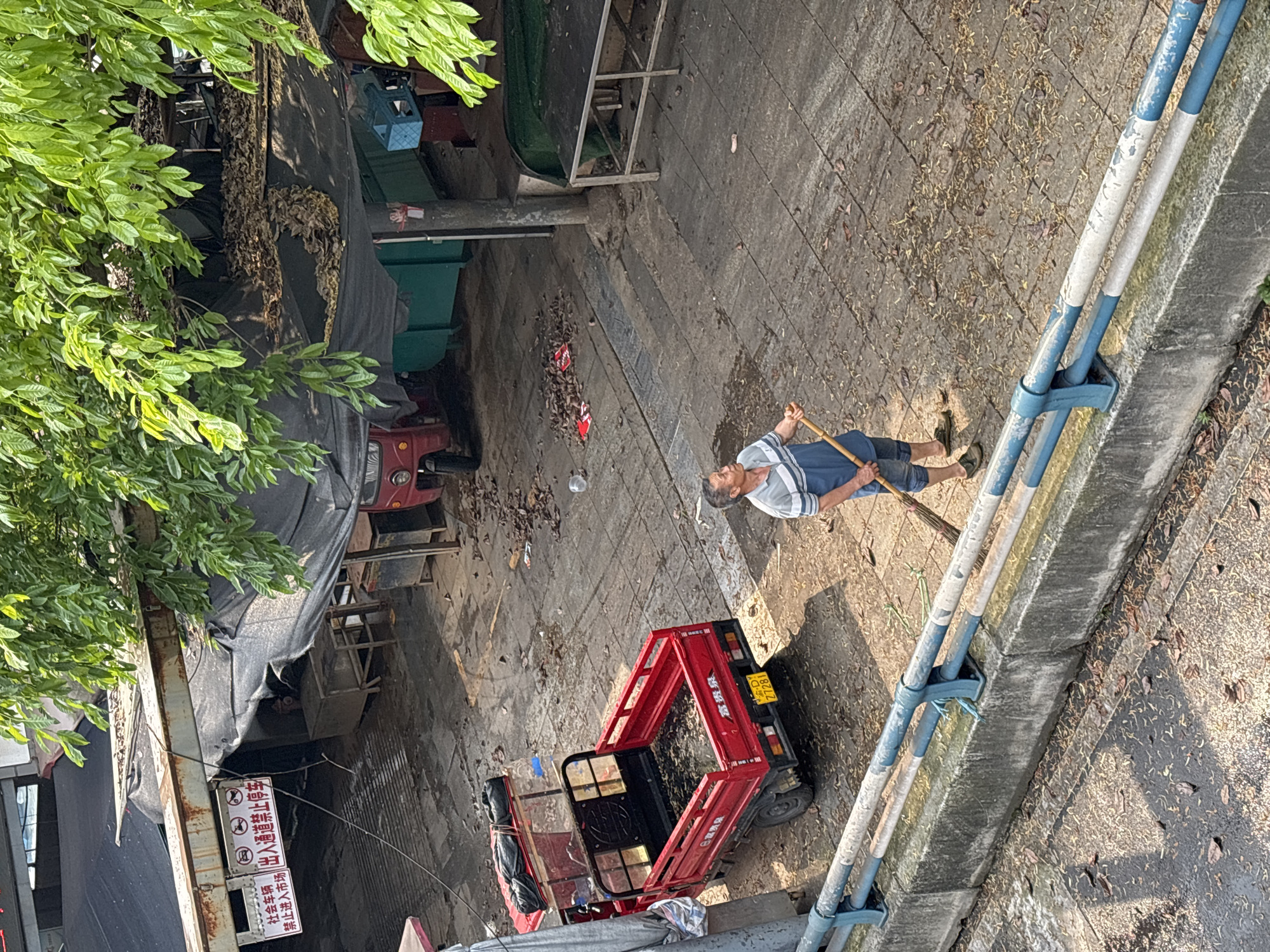 The image shows an aerial view of a street in Jiangjin, China. An elderly man wearing a striped shirt and blue pants is sweeping debris with a broom on a paved surface. Beside him is a red motorized tricycle parked on the street. Overhead structures are covered with black tarps. A metal railing with blue and white accents lines the right side of the street. A sign with Chinese characters is visible near the building, likely indicating local regulations. Green foliage overhangs the scene. The ground is littered with leaves and debris.