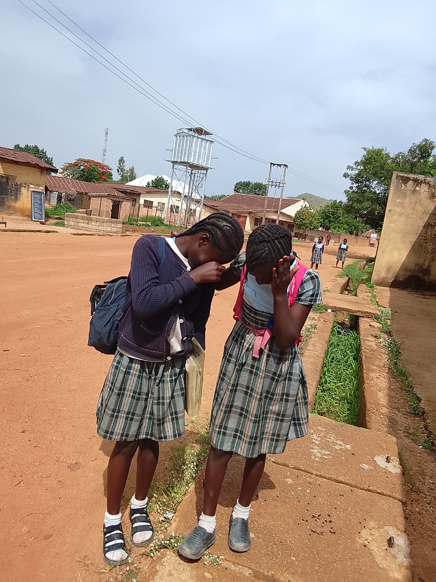 Two young girls stand close together outside a building, partially obscuring their faces with their hands. Both girls are wearing matching plaid school skirts, white socks, and dark shoes. The girl on the left has on black sandals and a navy blue cardigan, with a blue backpack slung over her shoulder, and the girl on the right has on gray shoes and a pink backpack. They are standing on a concrete ledge next to a ditch filled with green vegetation. In the background, there is a red dirt ground, buildings, and a water tower. In the distance, several people are walking away. The location appears to be a village or town in Unknown, Unknown.