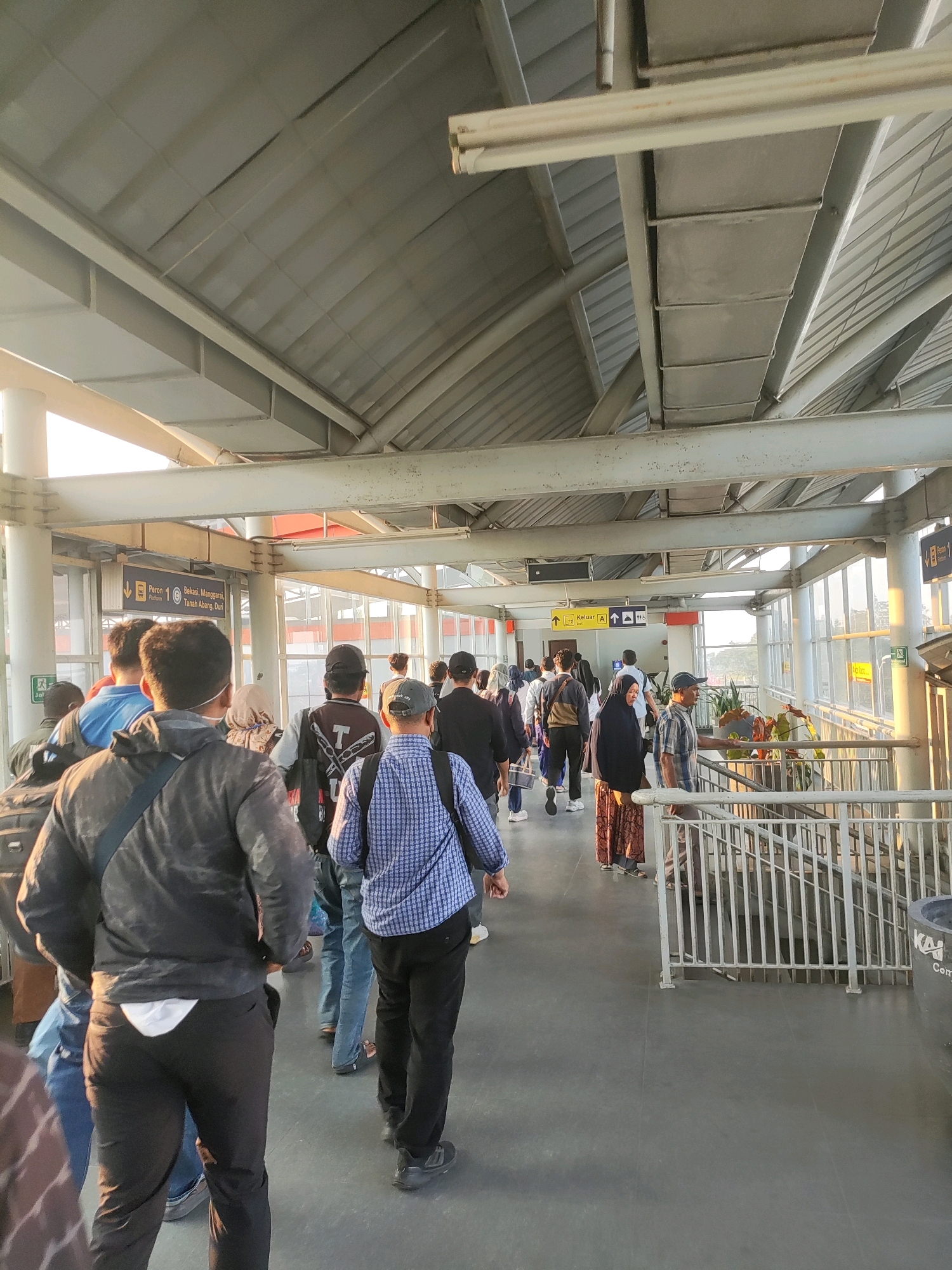 The image depicts an outdoor pedestrian walkway at a train station, likely within Telagamurni, Indonesia. A group of people are walking along the path, some in masks, moving towards the right side of the frame. The walkway is covered by a corrugated metal roof supported by industrial beams. To the left, a sign indicates "Peron 10" (Platform 10) with destinations including "Bekasi, Manggarai, Tanah Abang, Duri". Further down the walkway, another sign points towards an exit ("Keluar A"). The scene is illuminated by natural sunlight, casting shadows and highlighting the metallic structure of the walkway. A railing lines the right side of the path, with a staircase descending behind it. The ground is paved with a dark, smooth material. A person in the foreground on the left is wearing a dark jacket and black pants, while a person in the center wears a blue checkered shirt and dark pants.