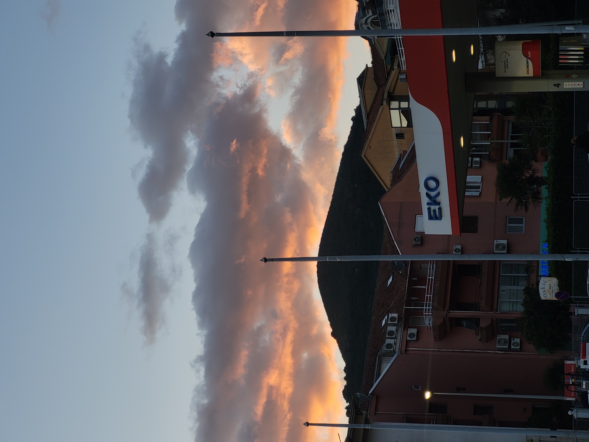 The image depicts a street scene in Budva, Montenegro, featuring an EKO gas station and surrounding buildings. The sky is visible with a sunset illuminating the clouds with orange and grey hues. A dark, tree-covered hill forms a backdrop to the buildings. Streetlights are visible in the foreground. The gas station, marked with the "EKO" logo, is adjacent to several multi-story buildings with numerous air conditioning units. There is a tree in front of the building on the right.