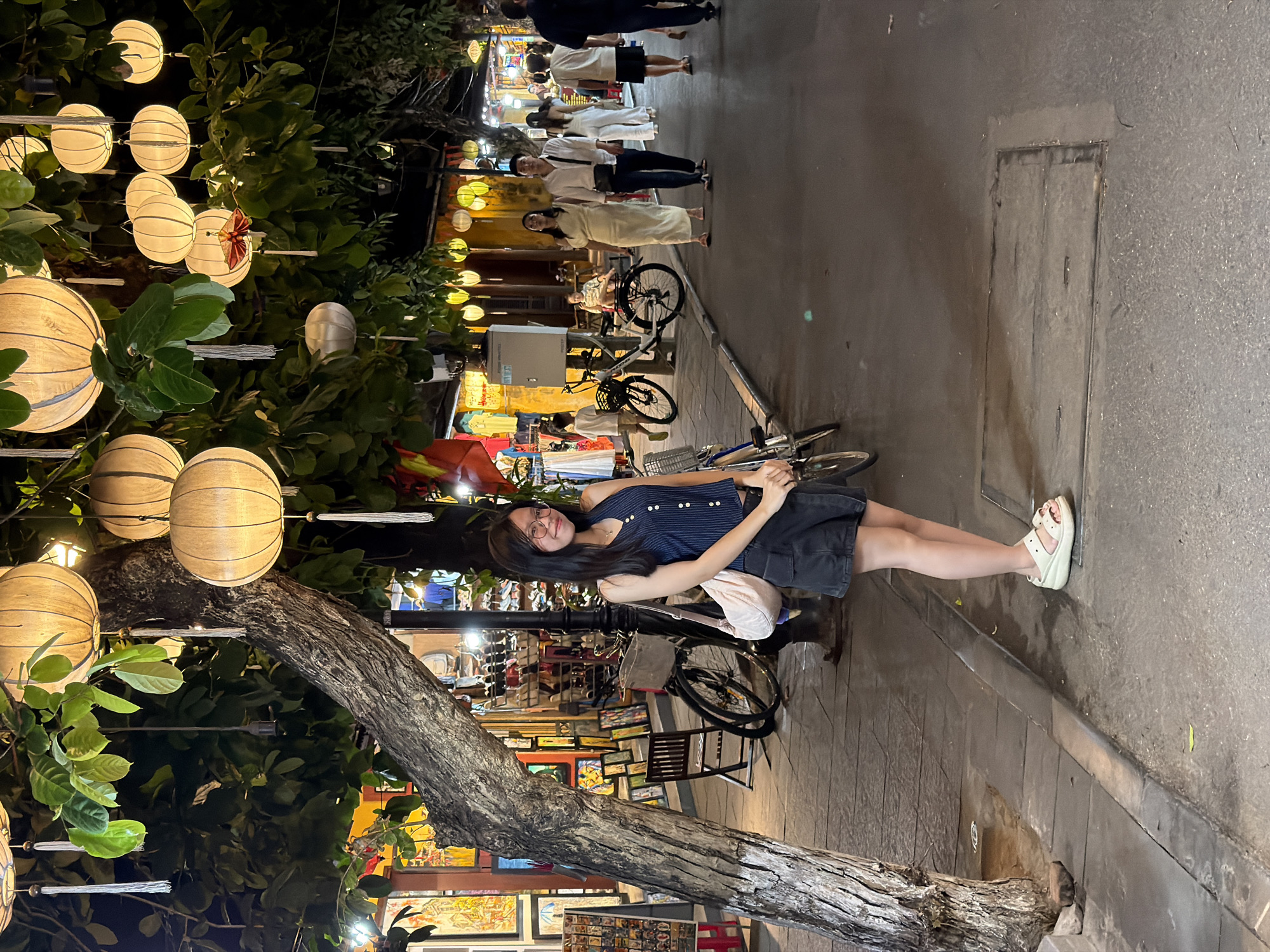 This is a nighttime street scene in Hoi An, Vietnam, with a young woman posing for the camera. She is wearing a dark, striped sleeveless top and a dark skirt, along with light-colored platform sandals. Her hair is dark and long, and she is wearing glasses. She has a light-colored bag slung over her shoulder.
The setting is a pedestrian street lined with trees adorned with decorative lanterns that cast a warm glow. The lanterns appear to be made of paper and are strung up on wires. In the background, there are shops and stalls, some displaying goods like artwork. Several bicycles are parked along the street. Other people are visible in the background, some walking and others standing.
The time of day is clearly evening or night, given the artificial lighting from the lanterns and shop signs. The atmosphere appears festive and vibrant, typical of Hoi An's ancient town area, which is known for its night markets and illuminated streets. The woman's pose and smile suggest a relaxed and happy demeanor. The overall mood is one of enjoyment and exploration of a charming, well-lit tourist destination. There is no visible text in the image that can be readily deciphered.