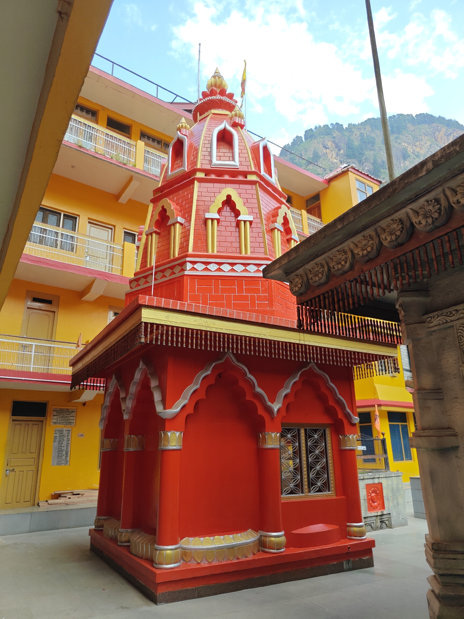 The image captures a vibrant red temple, likely Hindu, situated in a religious complex within Manikaran, India. The temple features a striking red facade with gold and silver embellishments, including a multi-tiered dome with arched decorative elements, a golden finial, and a small flag. Its base is adorned with arched alcoves and small pillars, all painted in the same bright red, gold, and silver palette.
In the background, a multi-story building painted a distinct bright yellow stands, equipped with numerous balconies featuring white railings. Windows and doors are visible across its various levels. Beyond these structures, a large mountain range, partially covered with trees, rises under a clear blue sky with scattered white clouds, suggesting a sunny daytime setting.
To the right, a detailed, carved stone or wooden structure with intricate floral patterns is partially visible. On the yellow building, a white sign containing text in Hindi is clearly discernible. The text reads: "श्री गुरुद्वारा श्री गुरू नानक देव जी मानिकरण साहिब" (Shri Gurudwara Shri Guru Nanak Dev Ji Manikaran Sahib) and "गुरुद्वारा समिति मानिकरण साहिब कुल्लू" (Gurudwara Committee Manikaran Sahib Kullu), followed by a list of names. This explicitly identifies a nearby Sikh Gurudwara and confirms the location as Manikaran Sahib, a significant pilgrimage site in Kullu, India. The scene is devoid of human activity, presenting a peaceful view of the architecture and natural environment.