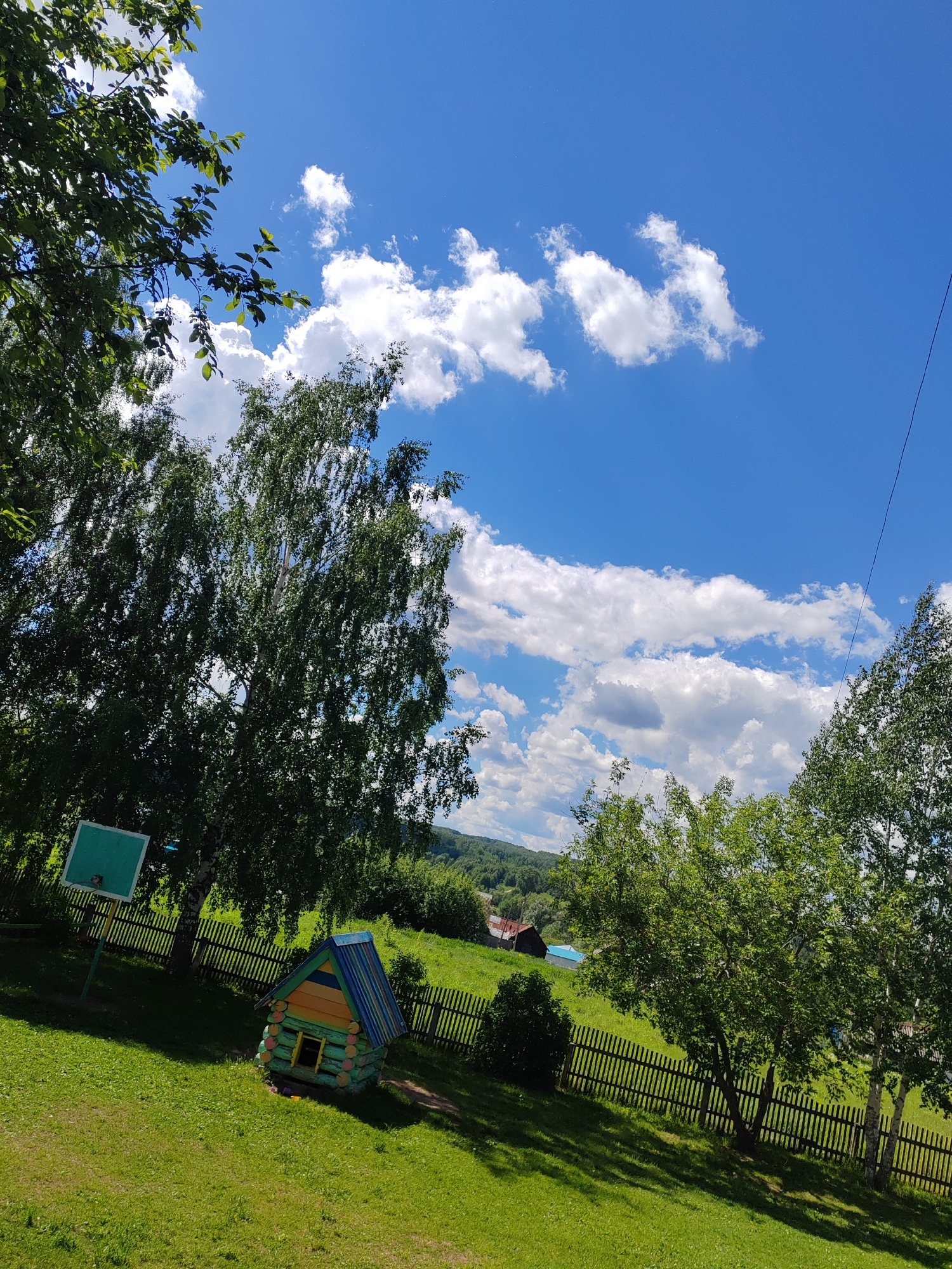 The image presents a sunny outdoor scene, likely in a rural or suburban setting. The sky is bright blue with scattered white clouds. The foreground features a grassy hill, with a small, colorful wooden playhouse. A wooden fence runs along part of the hill's slope. Trees are present on both sides of the frame, some obscuring the view of the distant landscape. In the background, there is a suggestion of hills and what appears to be a building with a red roof. The scene is peaceful and suggests a recreational or residential area.