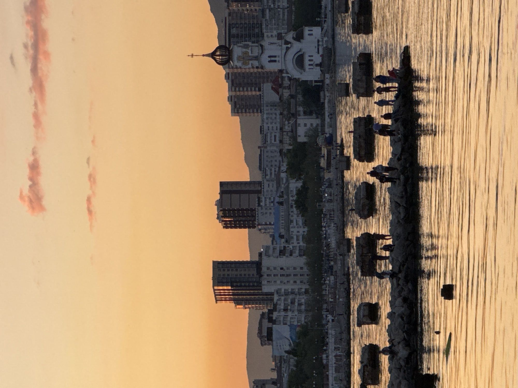 The image depicts a serene coastal cityscape at sunset or sunrise, likely Novorossiysk, Russia. In the foreground, a calm body of water, presumably the Black Sea, reflects the warm, golden light of the sky. Along the water's edge, a dark, rocky breakwater or shoreline is visible, upon which several people are standing. These figures appear as dark silhouettes against the brightly lit water, and their reflections are subtly cast onto the water's surface.
The midground showcases a dense urban skyline composed of various buildings, ranging from multi-story residences to commercial structures. A prominent feature within this cityscape is a white Orthodox church, distinguished by its classical dome, a golden cupola, and a cross at its apex. Behind the city, a line of mountains or hills forms a gentle silhouette against the horizon.
The background is dominated by the sky, displaying a beautiful gradient of warm orange and yellow tones, characteristic of the "golden hour." Faint, reddish, wispy clouds are scattered across the sky, adding depth and texture. The overall impression is one of peaceful observation, with the people seemingly enjoying the scenic view during this tranquil time of day. No visible text is present in the image.