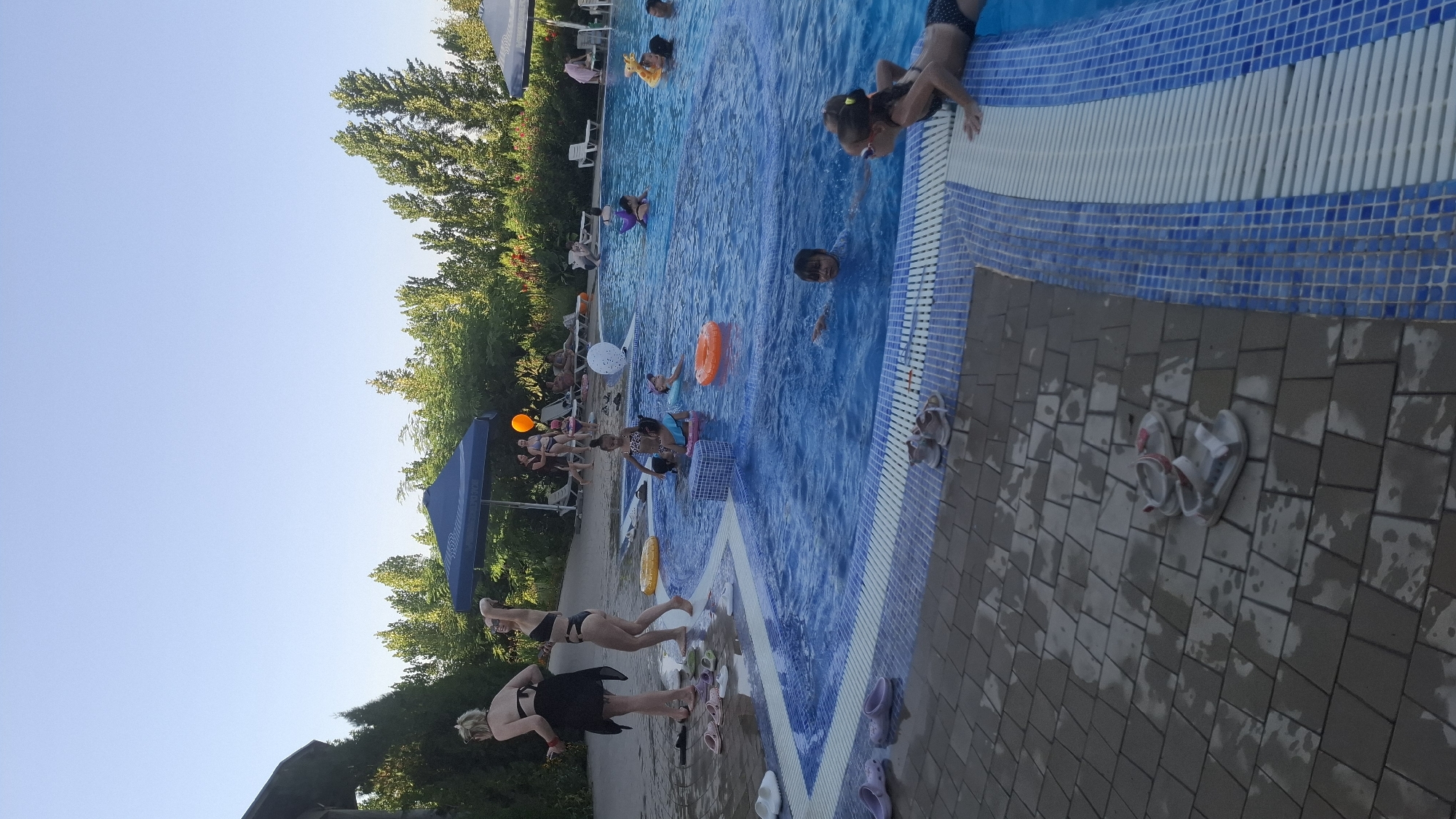 The image depicts an outdoor swimming pool area with several people, primarily children and adults, engaging in recreational activities. The pool is filled with clear blue water, and the surrounding area is paved with interlocking bricks and tiled borders.
In the foreground, on the tiled edge of the pool, a young girl in a dark swimsuit is partially visible, resting her arms on the pool's edge and looking towards the water. Further to the left, on the brick paving, there are several pairs of sandals and flip-flops, some appearing wet.
The pool itself contains multiple individuals. Several children are playing in the water, some with flotation devices such as inflatable rings. A few adults are also present in or near the water. In the background, there are lounge chairs and umbrellas, suggesting a public or resort-like setting. Trees and other vegetation are visible beyond the pool area, indicating an outdoor environment. The sky is clear and blue, suggesting fair weather.
While the image shows a recreational scene, the specific location within the city of Shymkent, Kazakhstan, cannot be determined solely from the visual information present in the photograph.