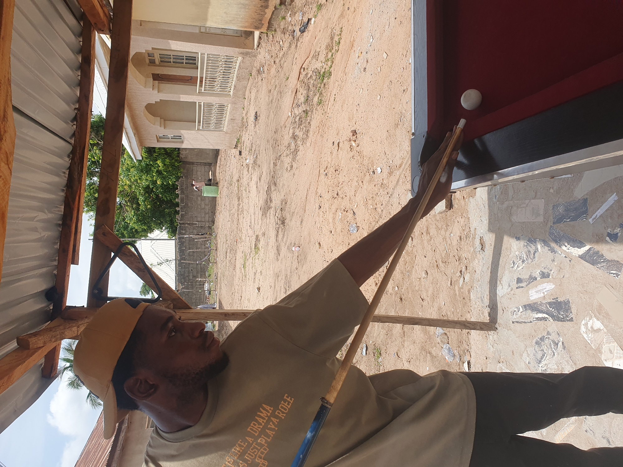 An adult male, wearing a tan baseball cap and a tan t-shirt with an orange graphic print, is playing billiards in an outdoor setting. He is positioned behind a red-felt billiards table, holding a cue stick and aiming at a white ball. The table appears to be set up under a makeshift wooden structure with a corrugated metal roof. The ground surrounding the table is sandy and uneven, with scattered debris. In the background, a light-colored building with arched windows and a white railing is visible, along with some trees and a fence. The overall environment suggests a rural or semi-urban area, possibly in Lokoja, Nigeria, given the architectural style and the natural elements present. The lighting indicates it is daytime with a bright sky, suggesting good weather conditions.