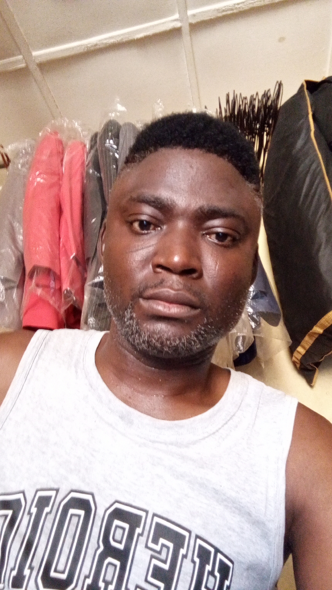 A medium close-up shot depicts a middle-aged Black man wearing a gray tank top with white lettering. He has a dark complexion, short black hair, and a sparse graying beard. His skin appears slightly moist. He looks directly at the camera with a neutral expression.
In the background are several pieces of clothing hanging on a rack, possibly in a shop or storage area. The clothes are covered with plastic bags. A red jacket is on the left and a black jacket with gold trim on the right. The location appears to be an indoor space, possibly a storage or retail environment in Sabon Tasha, Nigeria, although this cannot be determined definitively from the image alone.
