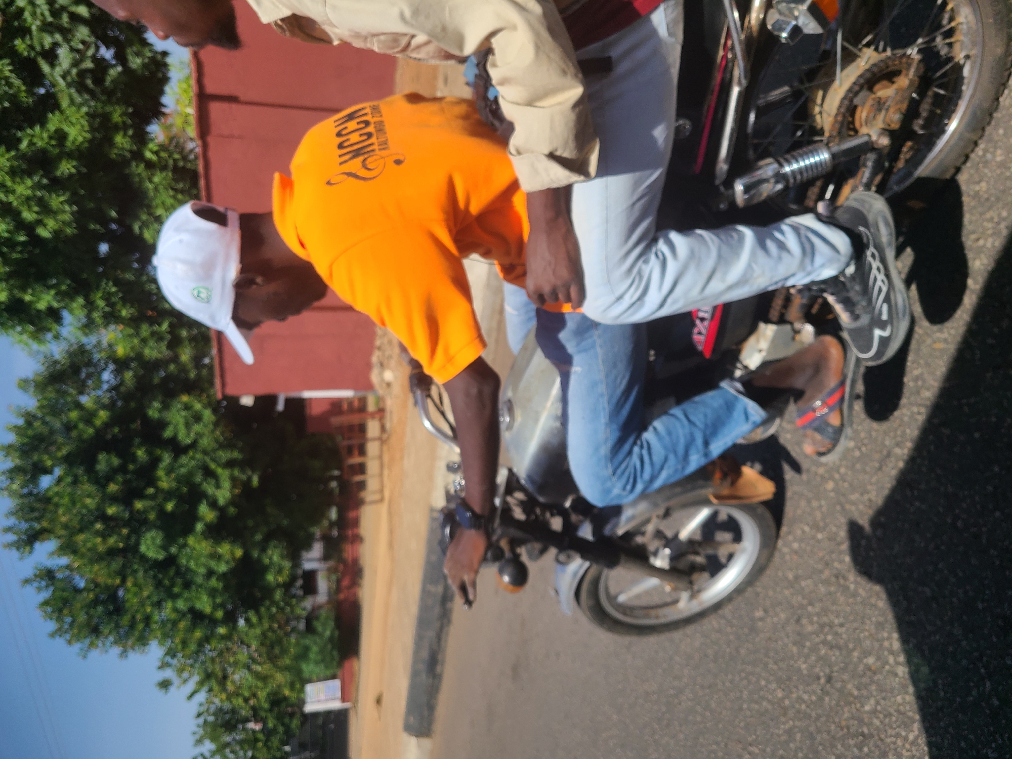 The image captures a motorcycle scene in Jos, Nigeria. Two individuals are on the motorcycle. The rider, wearing an orange t-shirt with the letters "NCCH" visible on the back, blue jeans, and a white baseball cap, is operating the vehicle. He is also wearing a wristwatch on his left wrist. The passenger is wearing a tan-colored shirt and light blue pants. The motorcycle is moving along a paved road, with trees and a red building in the background, typical of an urban landscape in Jos.