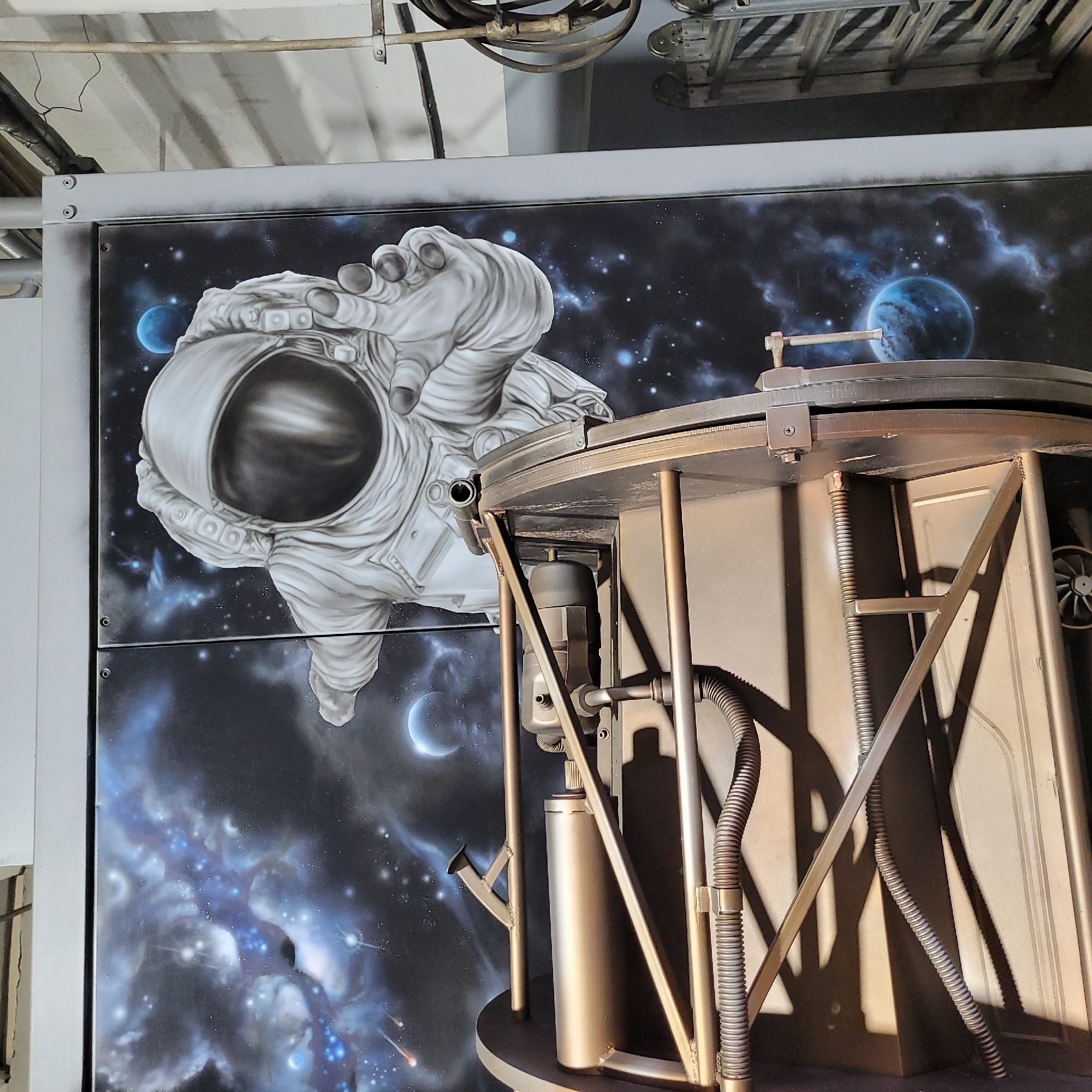 The media file displays an indoor scene featuring a prominent mural of an astronaut in space, partially obstructed by a metallic structure in the foreground.
The mural, rendered primarily in grayscale with a dark cosmic background, depicts an astronaut floating. The astronaut's right arm (viewer's left) is extended forward, with the hand seemingly reaching out. The helmet's visor is dark, reflecting the surrounding space. The background of the mural is a vibrant depiction of deep space, filled with countless stars, blue and purple nebulous clouds, a large blue planet, and a crescent moon.
In the foreground, a robust metallic structure, possibly made of bronze or dark-toned metal, stands. It features multiple vertical support rods, horizontal braces, and curved components, along with corrugated hoses or pipes. This structure is positioned in front of the mural, partially obscuring the lower right and central portions of the artwork. Its intricate framework casts distinct shadows on the mural behind it, indicating artificial lighting.
Above the mural and structure, industrial elements typical of a building's infrastructure are visible, including overhead pipes, conduits, and what appears to be a ceiling. The overall setting suggests a modern indoor space, possibly an exhibition area, a themed environment, or an industrial-chic establishment. There is no visible text in the image.