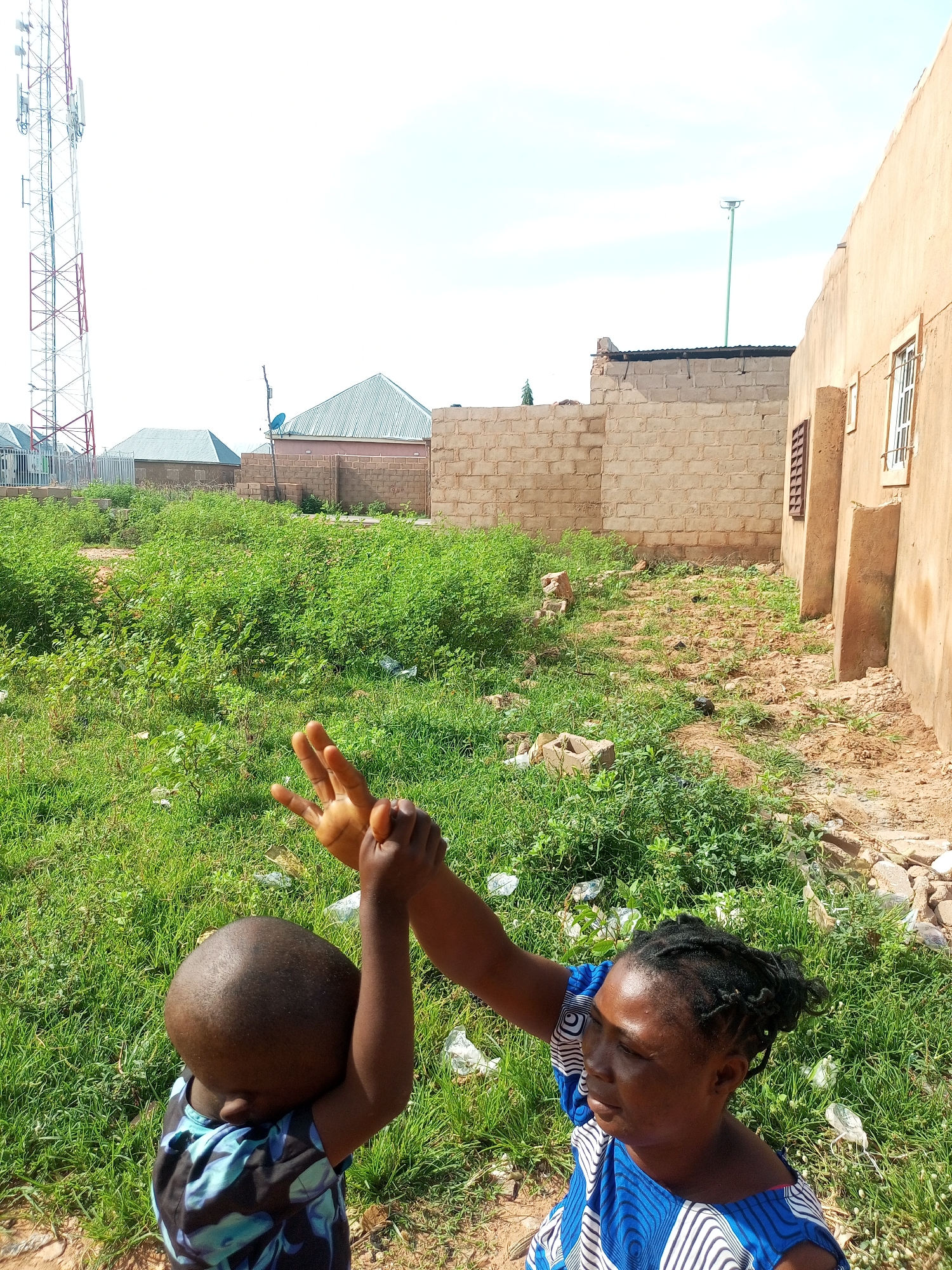 In Tempure, Nigeria, a medium shot captures a woman and a young child interacting outdoors. The woman, on the right, faces away from the camera with her head turned slightly to the left, her right arm extended upward and her hand forming a "peace" sign. Her blue and white patterned dress is visible. The child, on the left, faces away from the camera and appears to be reaching up towards the woman's hand, wearing a blue and black patterned shirt. They are standing in a patch of tall green grass, with some debris scattered around. In the background, there are several structures including a tall cell tower on the left, followed by various low-rise buildings with corrugated metal roofs. A light brown, unfinished brick wall runs along the right side of the frame, with part of a building with a window visible. The sky is bright and clear.