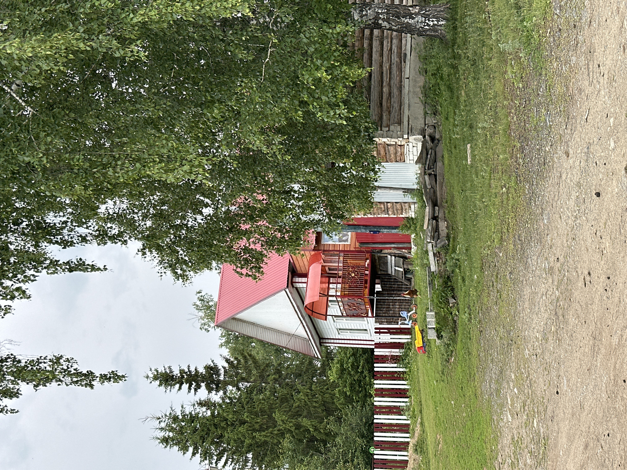 A rural scene is depicted under an overcast sky, with a white wooden house featuring a vibrant red roof and a red-canopied balcony. Adjacent to the house is a log structure. In the foreground, a gravel path leads to a grassy area with a red and white striped fence. Scattered on the grass are children's toys, including a yellow wagon and a blue tricycle. A chicken stands near the toys. Trees, including a large birch tree with peeling bark, frame the scene.
