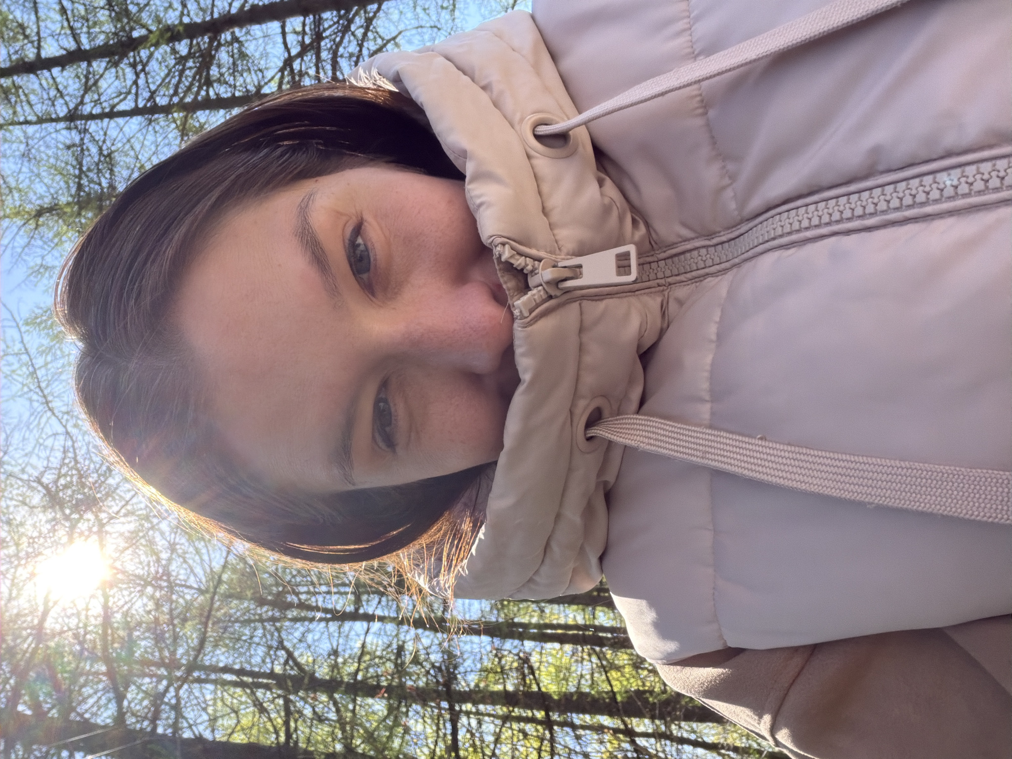 A young woman's face is angled towards the camera, with a pale skin tone and visible freckles. She has dark brown hair with sunlight filtering through some strands, highlighting them with a golden hue. She is wearing a light beige puffer jacket, zipped up to her neck, obscuring part of her face. The jacket hood is pulled around her head, and drawstrings are visible.
The background shows a partial view of trees and sky, with bright sunlight breaking through the branches. The location might be a park or wooded area, but there is no specific landmark or identifying feature to pinpoint it to Staraya, Russia.