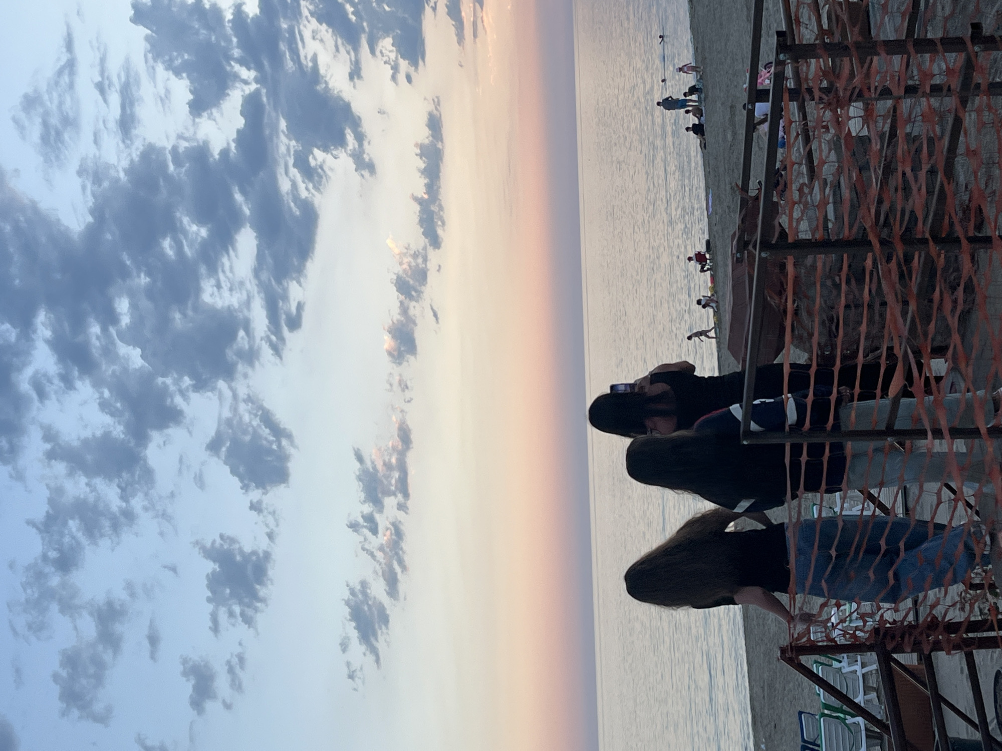The image shows a beach scene in Alanya, Turkey at sunset. The sky is filled with clouds on the left, transitioning to orange and pink hues near the horizon, and a calm sea stretches horizontally across the frame. In the foreground, three individuals, possibly women, are standing near a fenced-off area constructed with metal frames and orange plastic barriers. One person wears a black top and jeans, another has a black top with blue and white stripes, and the third wears a black top and blue jeans. People are scattered along the beach in the background, some near the water, some seated or standing. Beach chairs are visible near the bottom of the frame.