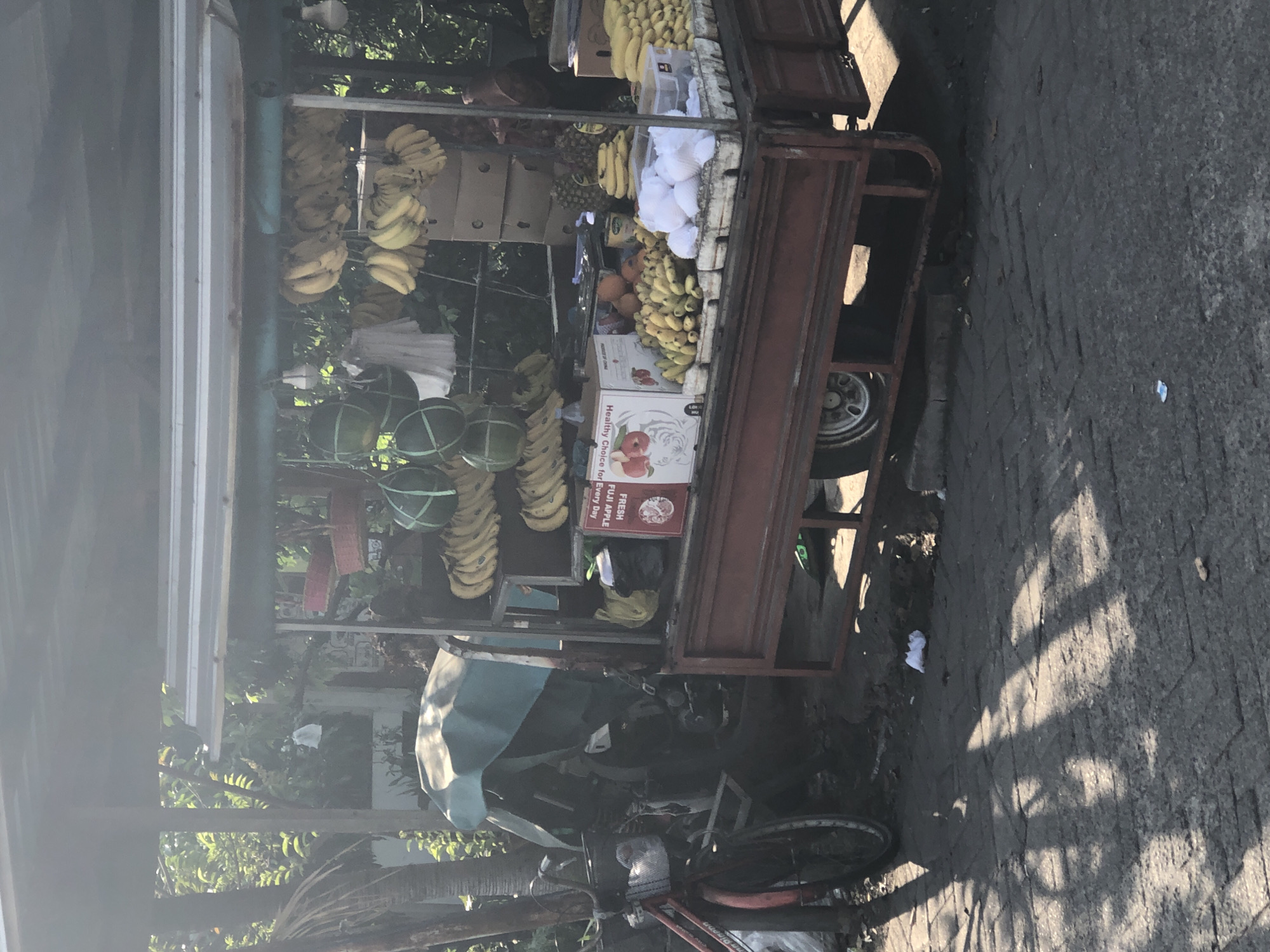 The image depicts an outdoor fruit stand in Balongdowo, Indonesia, with a tricycle-based cart laden with various fruits. The cart is covered by a corrugated metal roof and has a red painted body. Numerous bunches of bananas are hanging from the roof and displayed on shelves. Other fruits visible include watermelons, pineapples, and what appear to be oranges and smaller bananas. Cardboard boxes are stacked behind the fruit display. To the left of the cart, a red bicycle with a basket is parked, and a covered motorcycle is partially visible behind it. The ground is paved with interlocking bricks. Sunlight casts shadows across the scene, indicating it is daytime.