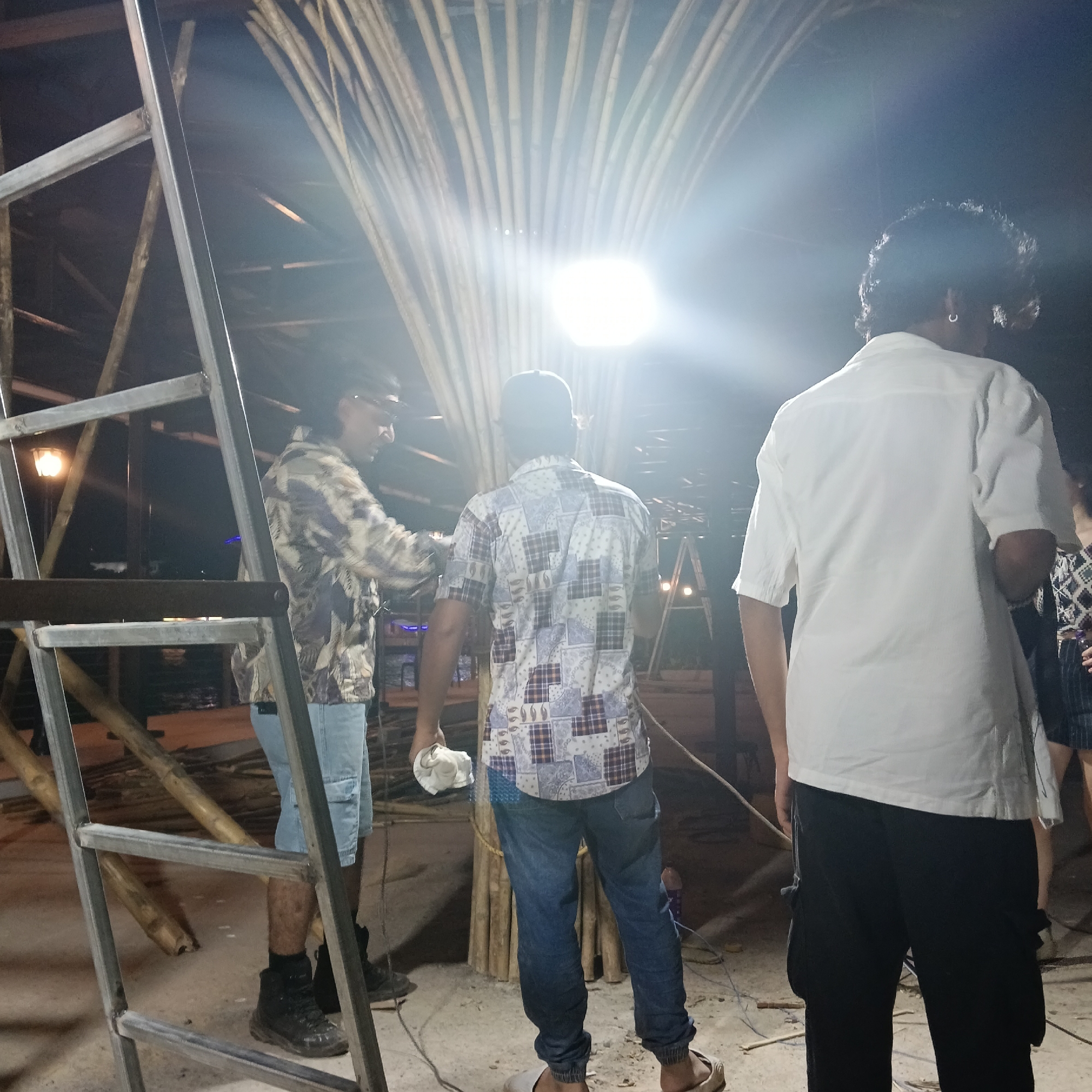 The image appears to have been taken at night in what might be Panaji, India, based on architectural styles and clothing. A metal ladder frame stands in the left foreground. Three men are present. The man on the left wears a camouflage shirt, blue shorts, and work boots. The central man is dressed in a patterned shirt and jeans with sandals. The man on the right wears a white shirt and dark pants. Behind them is a structure constructed of bundled bamboo poles, illuminated by a bright light source. Electrical cords lie on the ground.