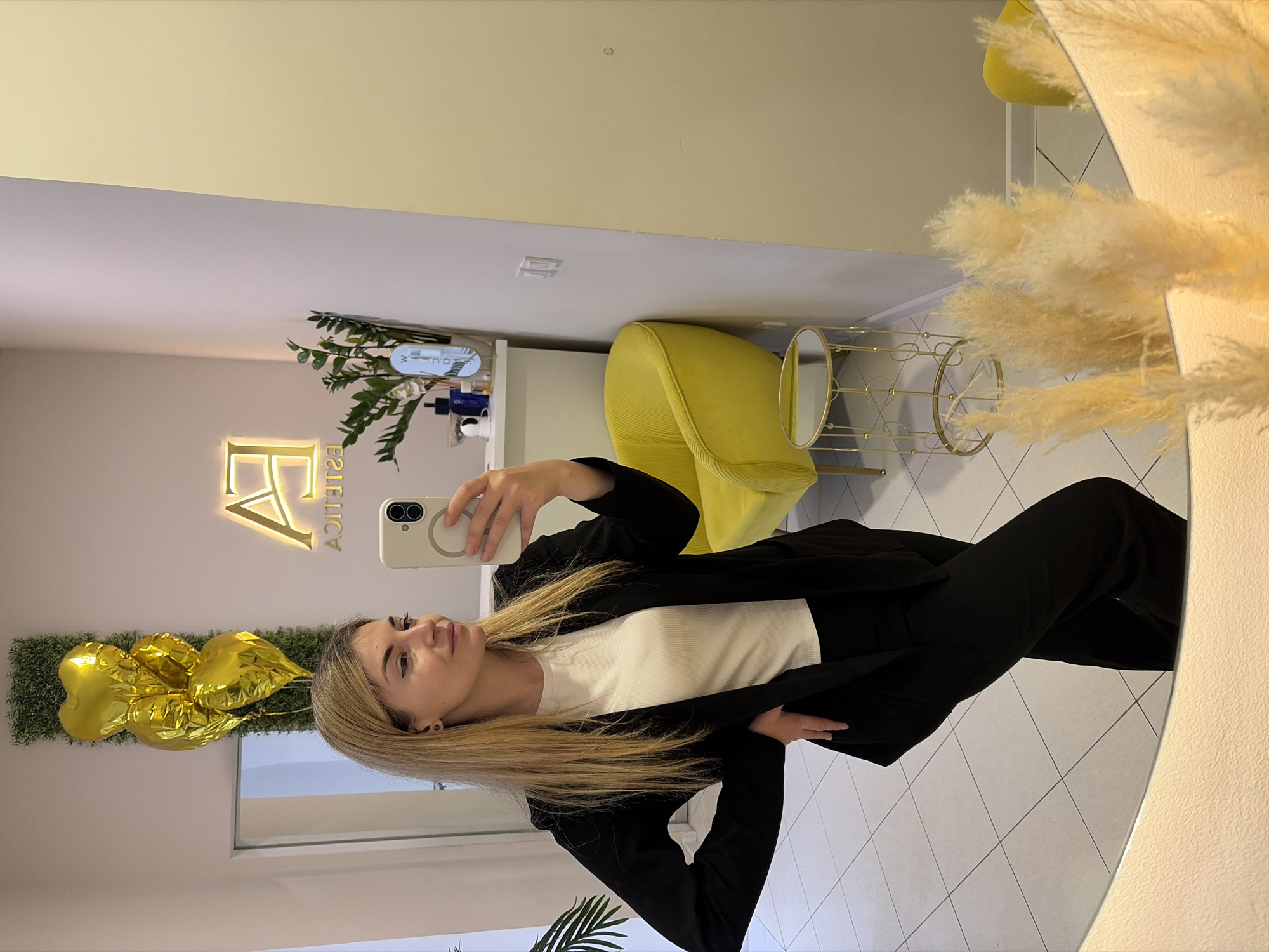 A woman is taking a selfie in a mirror. She is wearing a black suit and a white shirt. The background includes a yellow chair, a table, and a sign that says "FA Estetica". The location appears to be an interior space. The city of Budva, Montenegro is not discernible from this image.