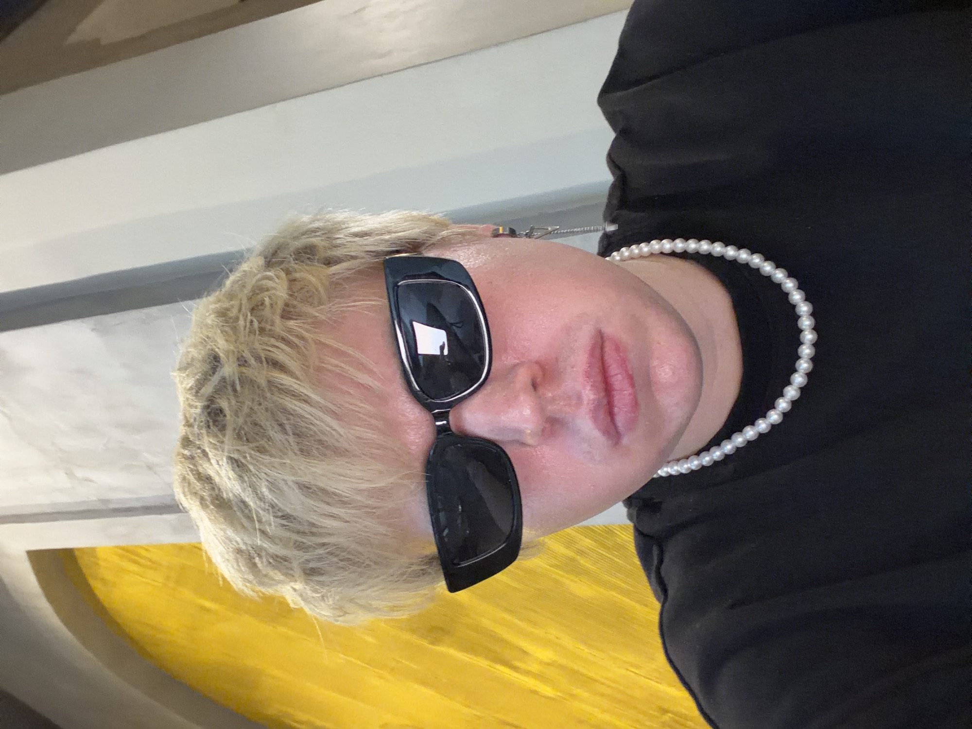 A close-up selfie-style portrait taken in Thủ Đức, Vietnam, features an individual with short, light blonde hair and a fair, somewhat flushed complexion. The person wears dark, rectangular sunglasses with a slight upward curve at the outer edges, obscuring their eyes. A necklace composed of white, spherical pearls is visible around their neck, paired with a plain black t-shirt. A silver-toned earring, consisting of a hoop from which a chain and a small geometric charm dangle, is worn on the subject's right ear. The background comprises a vibrant, textured yellow wall or architectural element with a curved upper edge on the left side of the frame. To the right, a light grey wall with subtle vertical darker grey stripes or panels is present.
