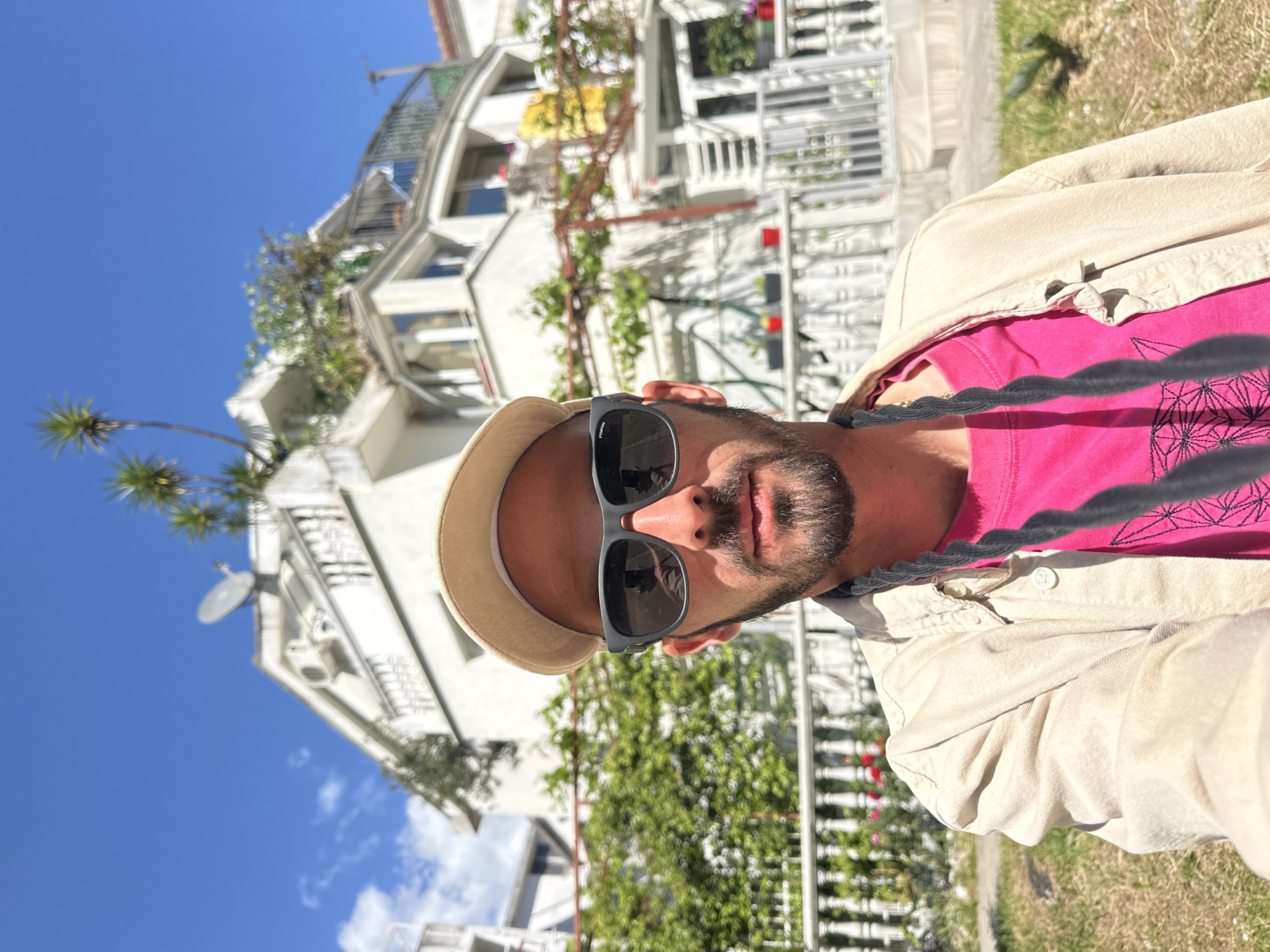 A selfie of a man wearing sunglasses and a hat, with a beard, pink shirt, and tan jacket, stands in front of a white building with plants and a blue sky. The scene is likely located in Budva, Montenegro.