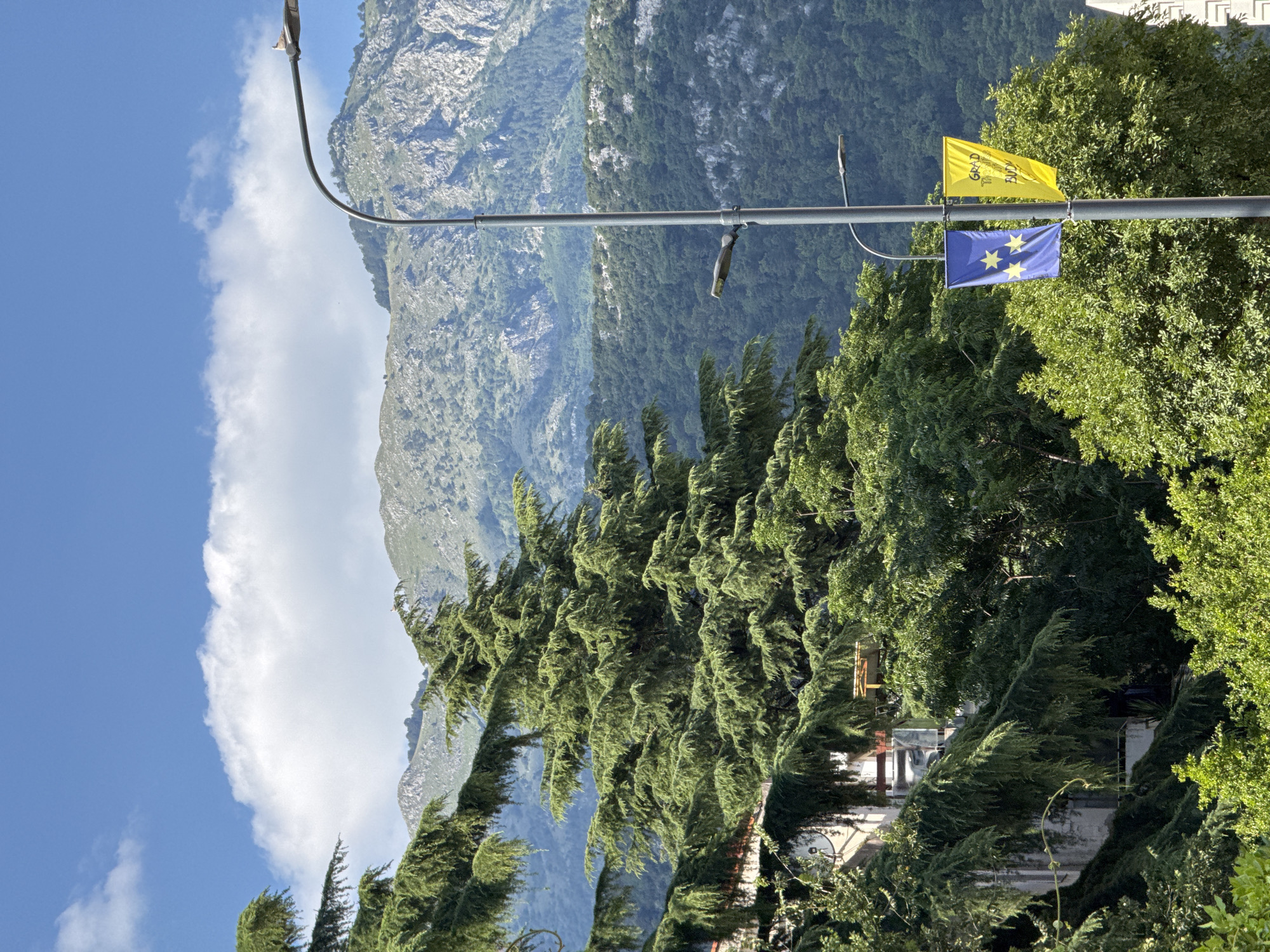 In Budva, Montenegro, a tall grey lamppost stands in the foreground against a backdrop of lush green trees and mountainous terrain. The lamppost is adorned with two vertical banners: one blue with yellow stars and another yellow banner with text that is partially obscured. In the upper portion of the image, a bright blue sky is dotted with fluffy white clouds, and the distant mountains are rugged and covered in green foliage. The scene appears to be an outdoor, daytime setting.