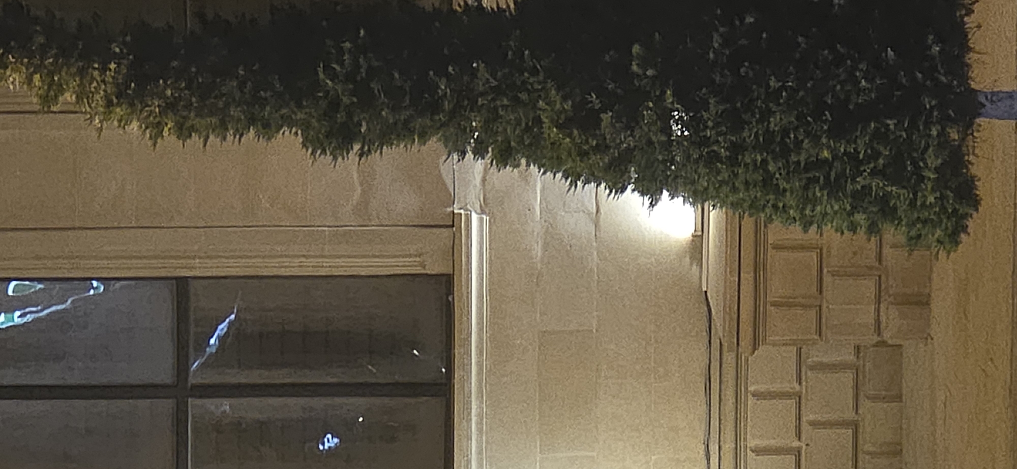 The image depicts a dark, outdoor scene with a building facade and a coniferous tree. The building features a large window with multiple panes, some of which appear to have reflections of blue light. Below the window, the facade is constructed of large, light-colored stone blocks, some of which are ornately carved. A light source, possibly a wall-mounted lamp, illuminates a section of the stone wall. The tree, which is dark green and conical in shape, is positioned in the foreground to the right of the building. The lighting suggests it is nighttime or dusk. The scene is likely a public space in Sumgayit, Azerbaijan, potentially near a historical building or a well-lit urban area. There are no people or animals visible in the image, and no discernable actions are taking place. The environment appears calm and still.