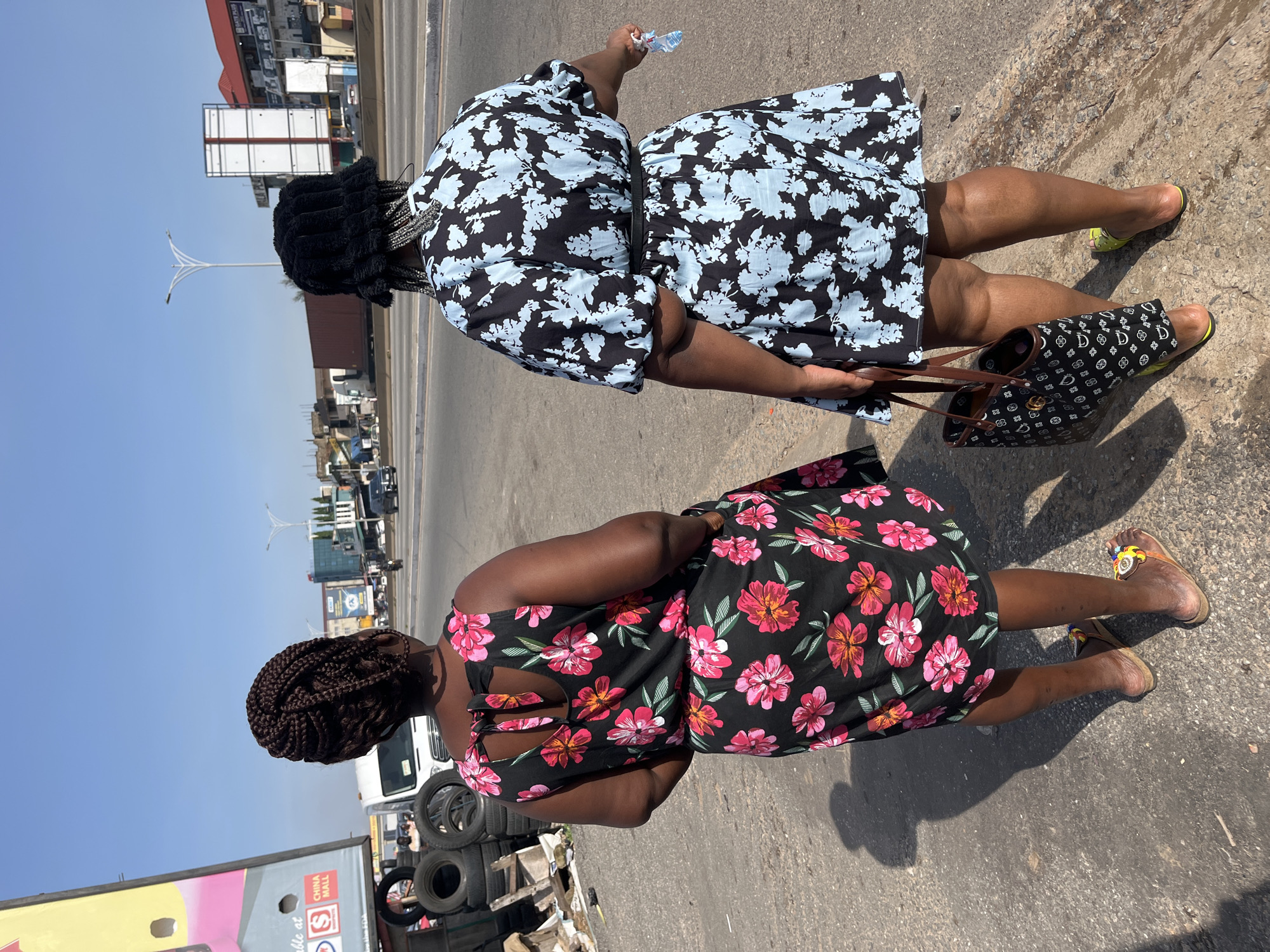 Two women stand on a paved roadside in Accra, Ghana, viewed from behind. The woman on the left wears a black sleeveless dress with a pink floral print and a bow tie closure at the back. She has braided hair. The woman on the right wears a black bucket hat and a black dress with a light blue floral pattern. She carries a patterned tote bag and a small plastic bottle in her right hand. The background shows a busy street with vehicles, buildings, and streetlights, indicative of an urban environment in Accra. There are stacks of tires and debris on the left side of the image, suggesting an informal commercial area. The sky is clear and blue.
